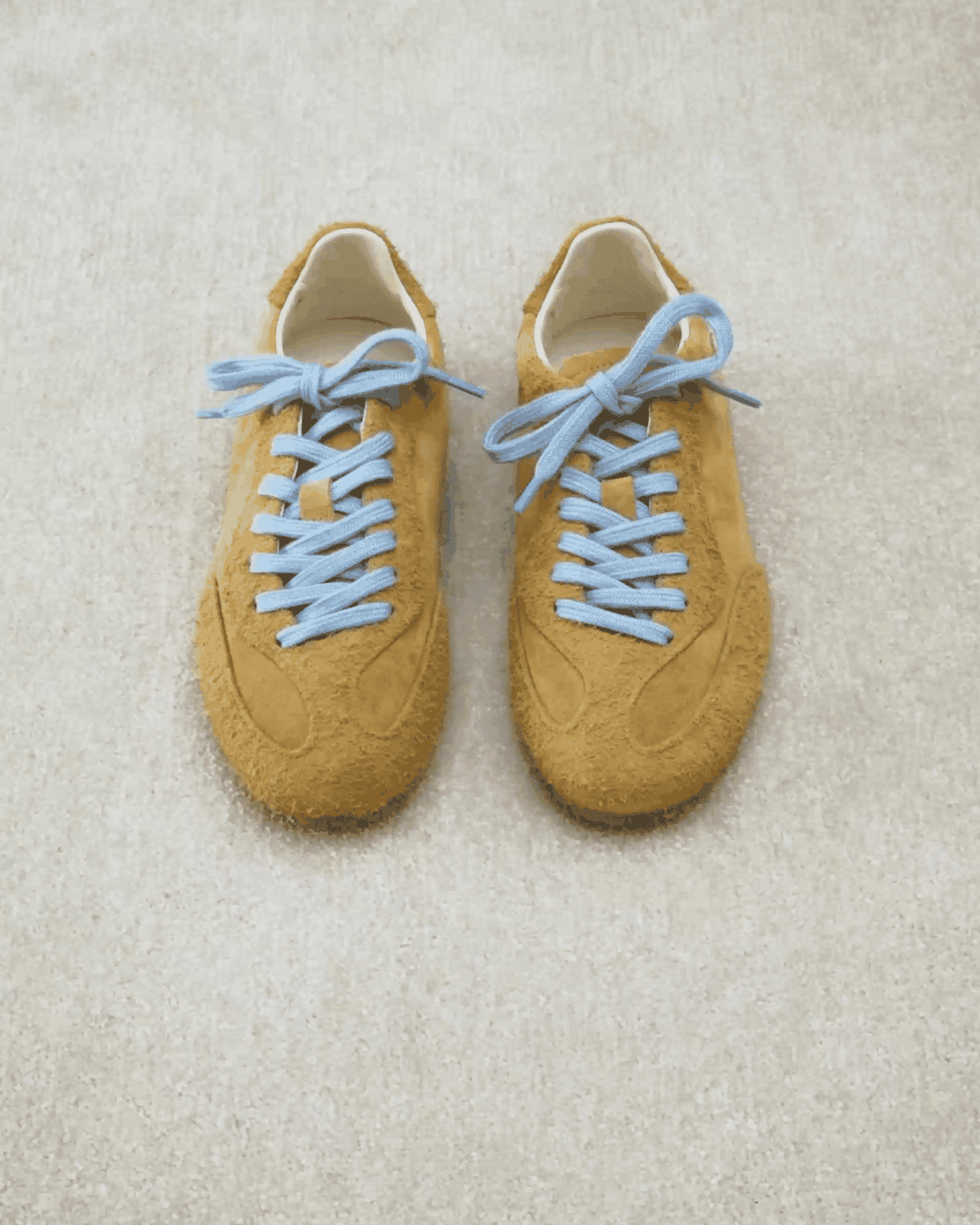Brown suede sneakers with light blue laces on a beige carpet.