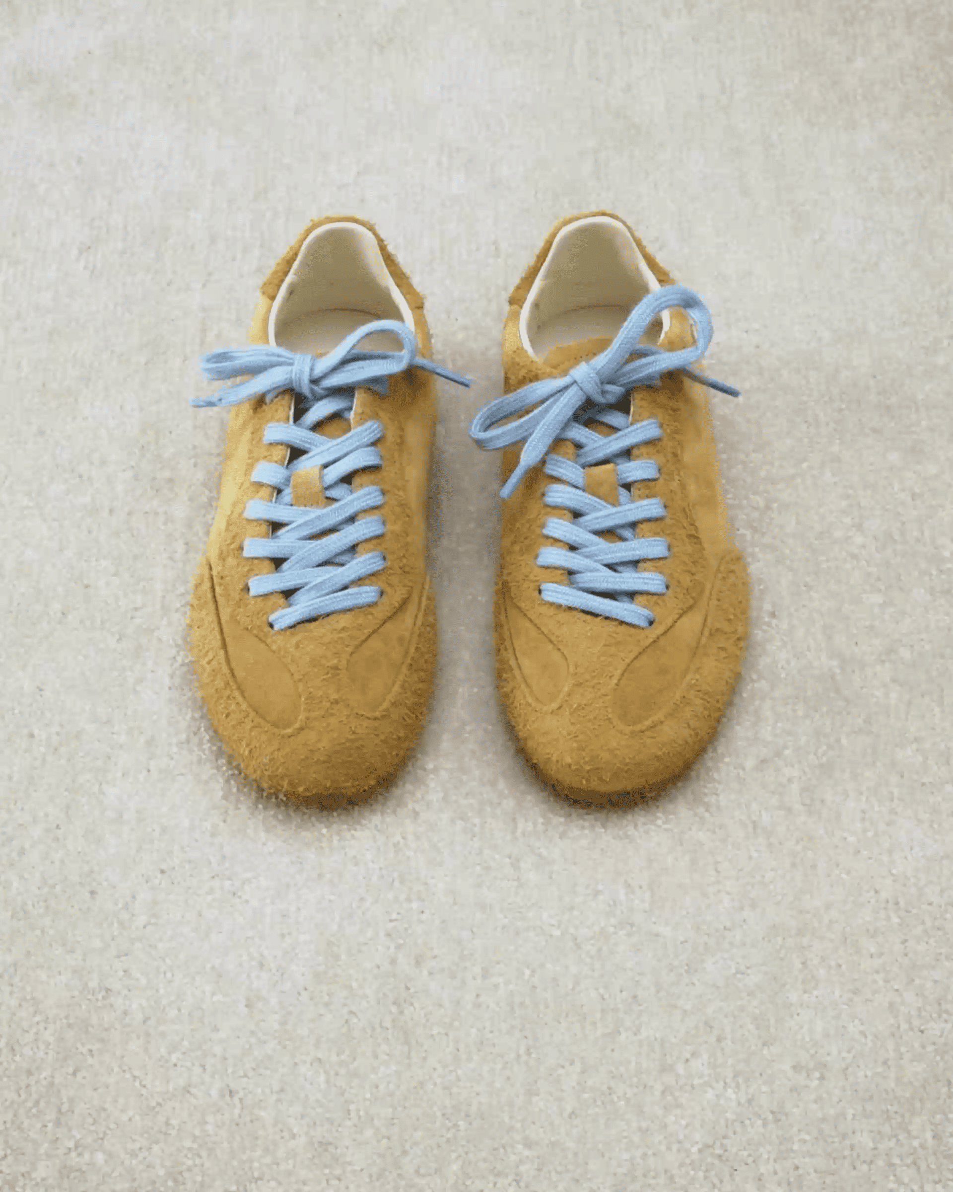Brown suede sneakers with light blue laces on a beige carpet.