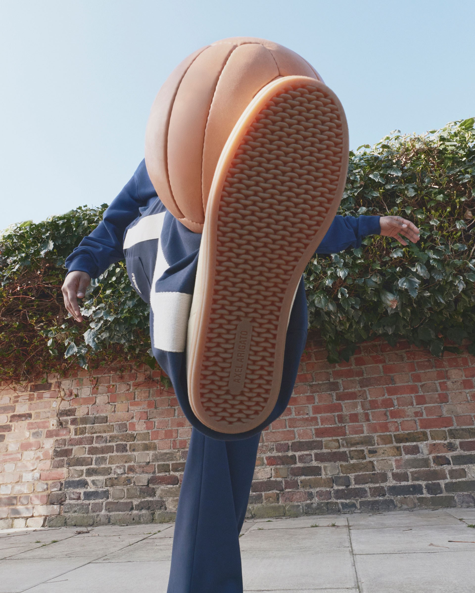 Low-angle view of a person in a tracksuit kicking a basketball with a textured sole of their shoe, against a brick wall and ivy background.