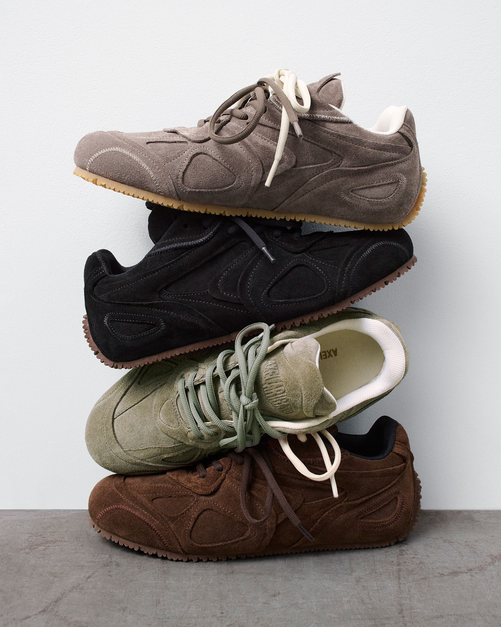 A stack of four suede sneakers in gray, black, green, and brown, with gum soles, against a plain background.