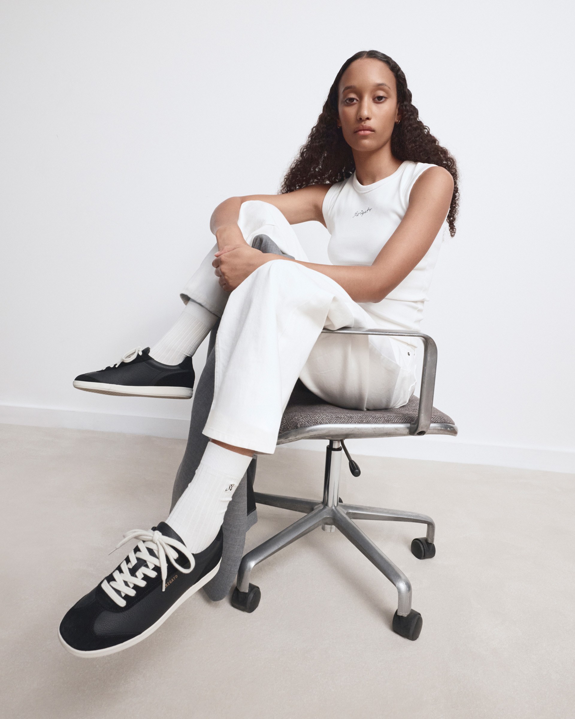 Person with long curly hair sits on a swivel chair wearing a white outfit and black sneakers, against a plain background.