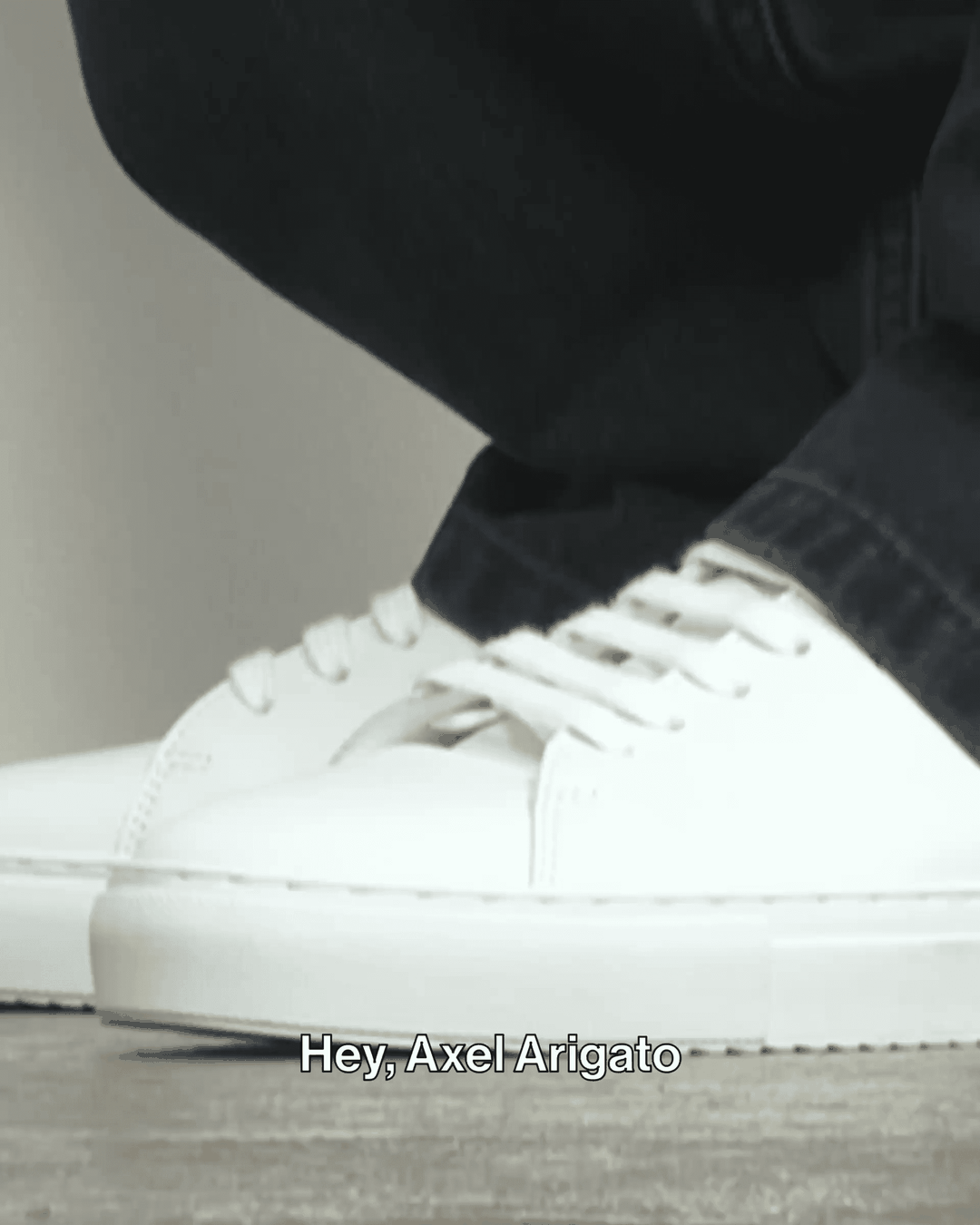 Close-up of white sneakers paired with black pants. Subtitles at the bottom say, "Hey, Axel Arigato."