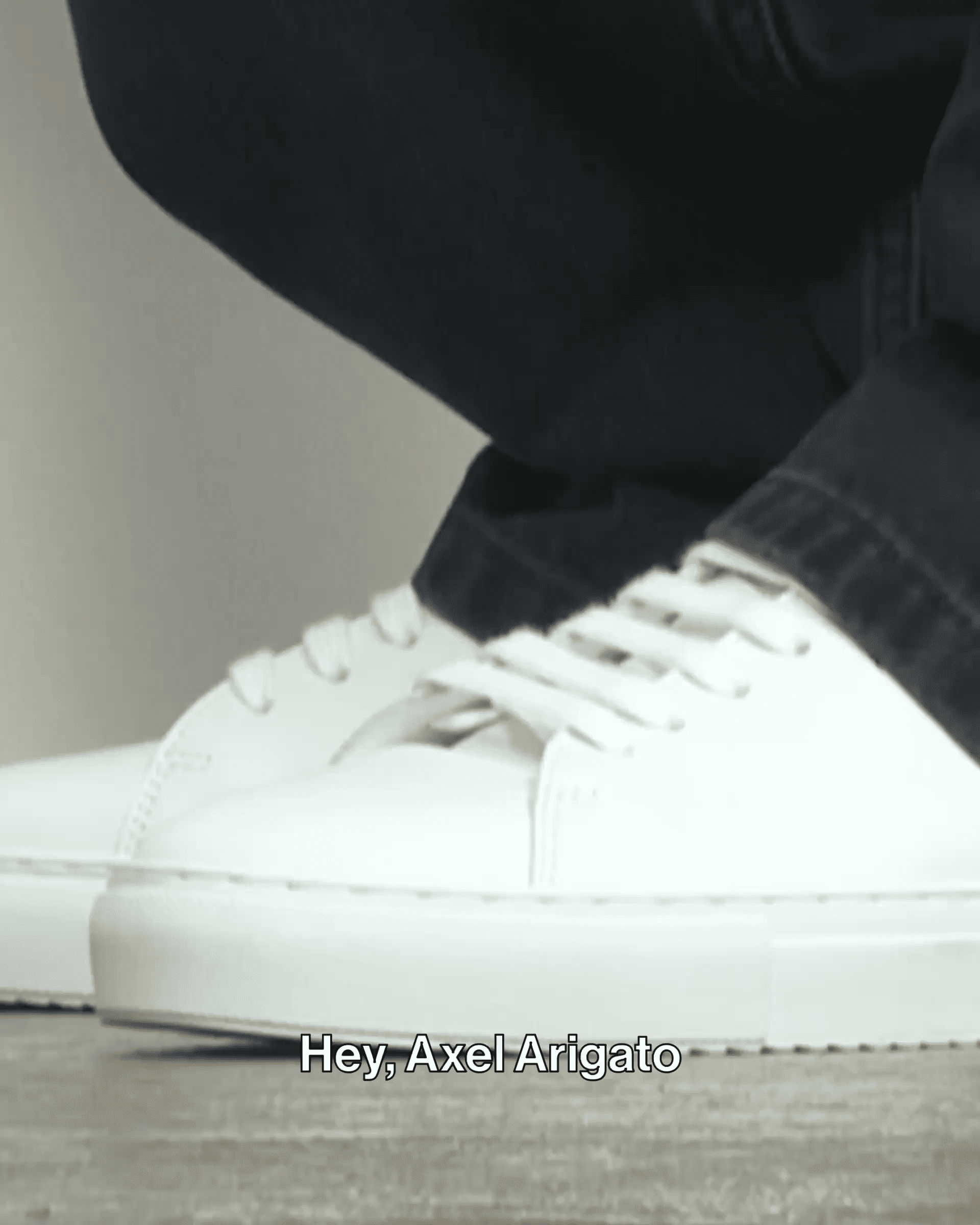 Close-up of white sneakers paired with black pants. Subtitles at the bottom say, "Hey, Axel Arigato."