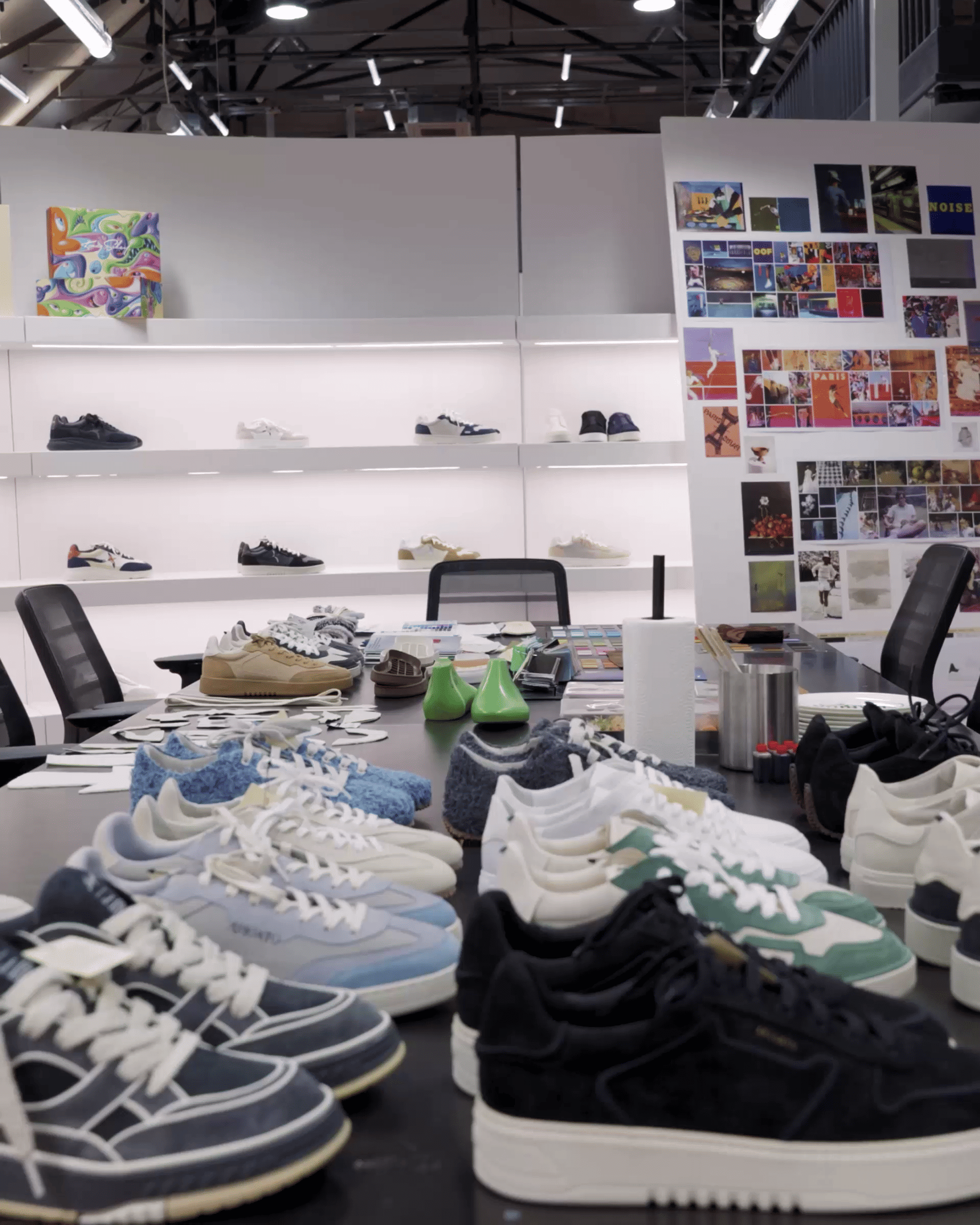 A designer showroom with various sneakers on a table and shelves, and a wall collage of photos and inspirations in the background.