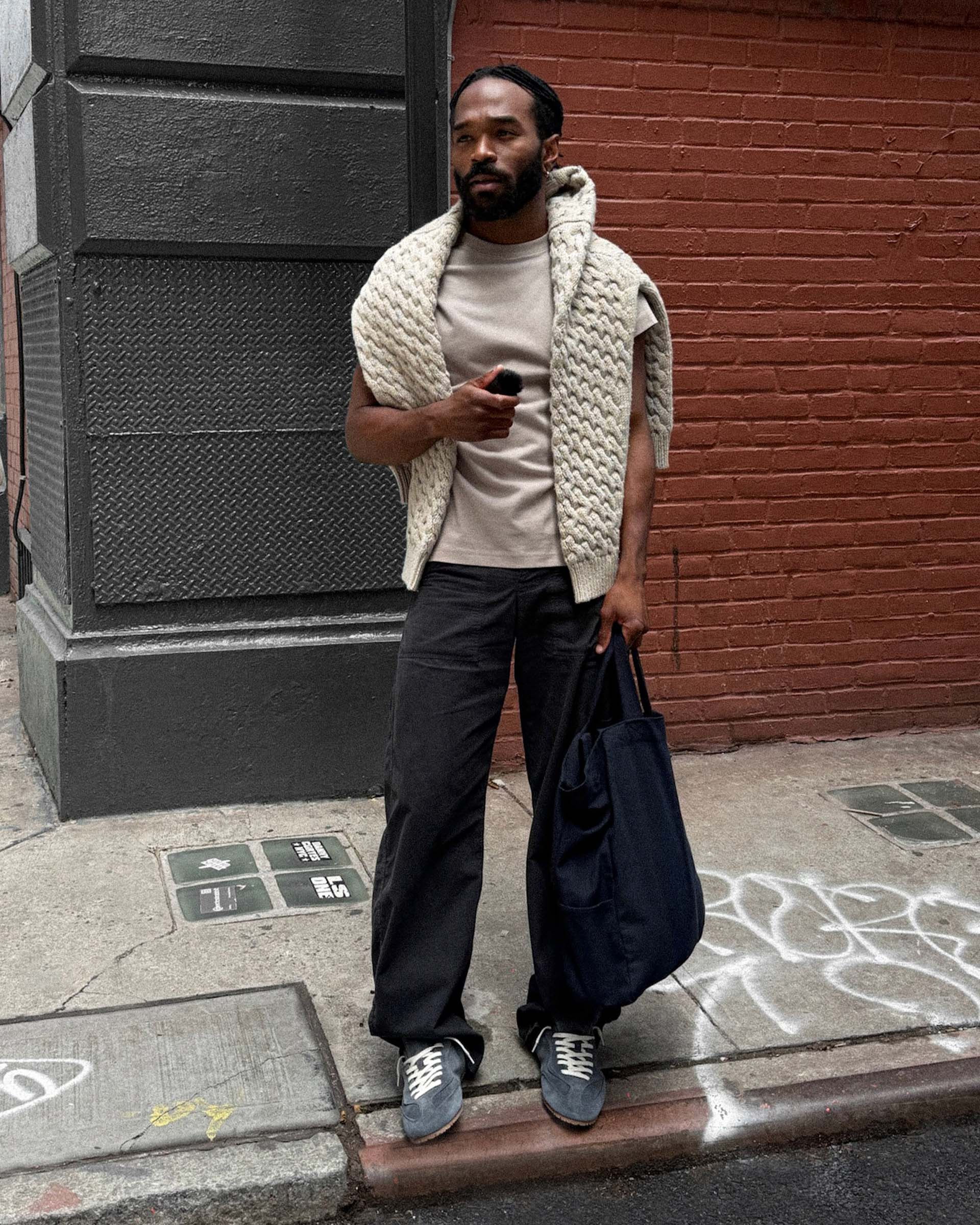 Man in casual attire with a knit sweater draped over shoulders, holding a phone and carrying a large bag, stands on a city sidewalk.