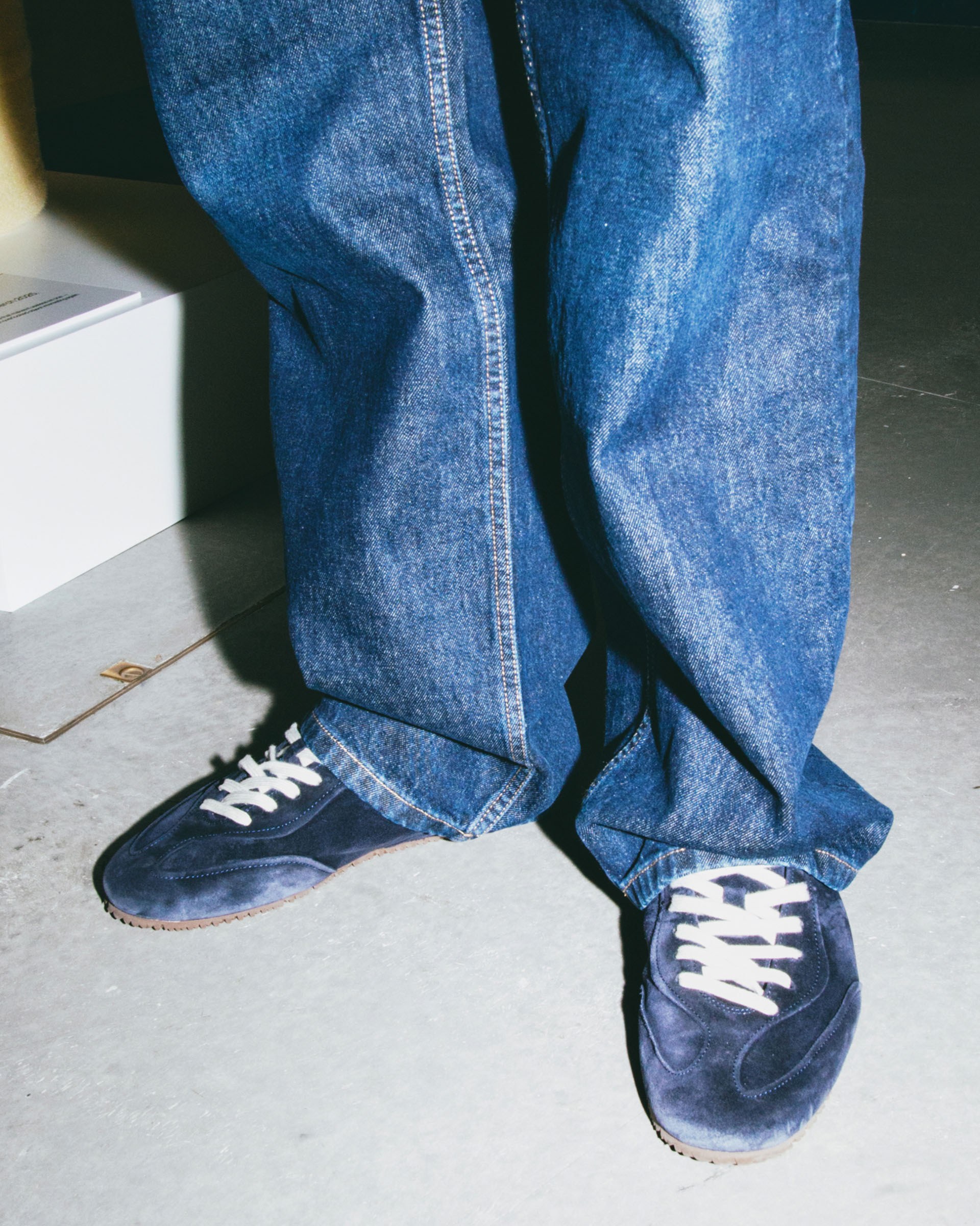 Person wearing baggy blue jeans and navy sneakers with white laces, standing on a gray floor.