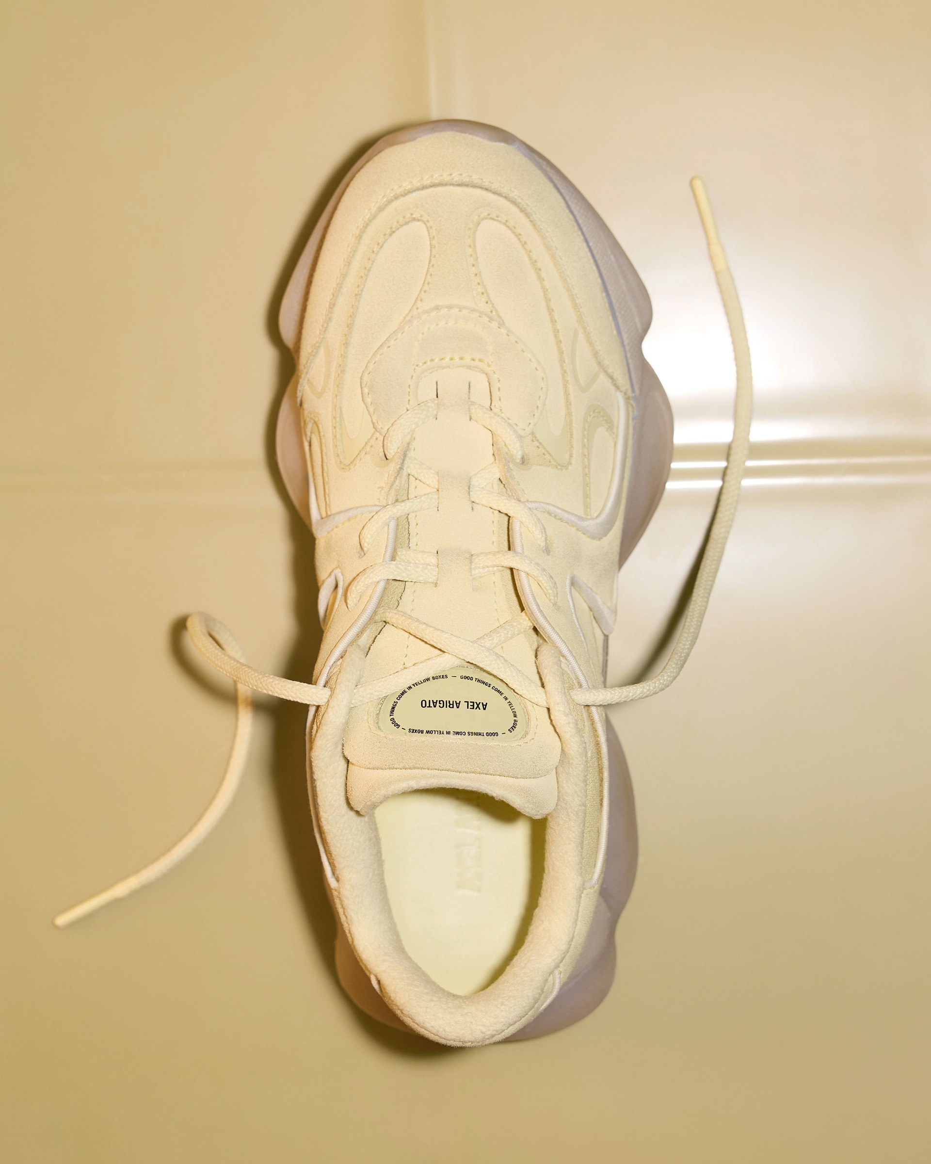 Top view of a single white sneaker with a chunky sole and untied laces on a light-colored tiled floor.