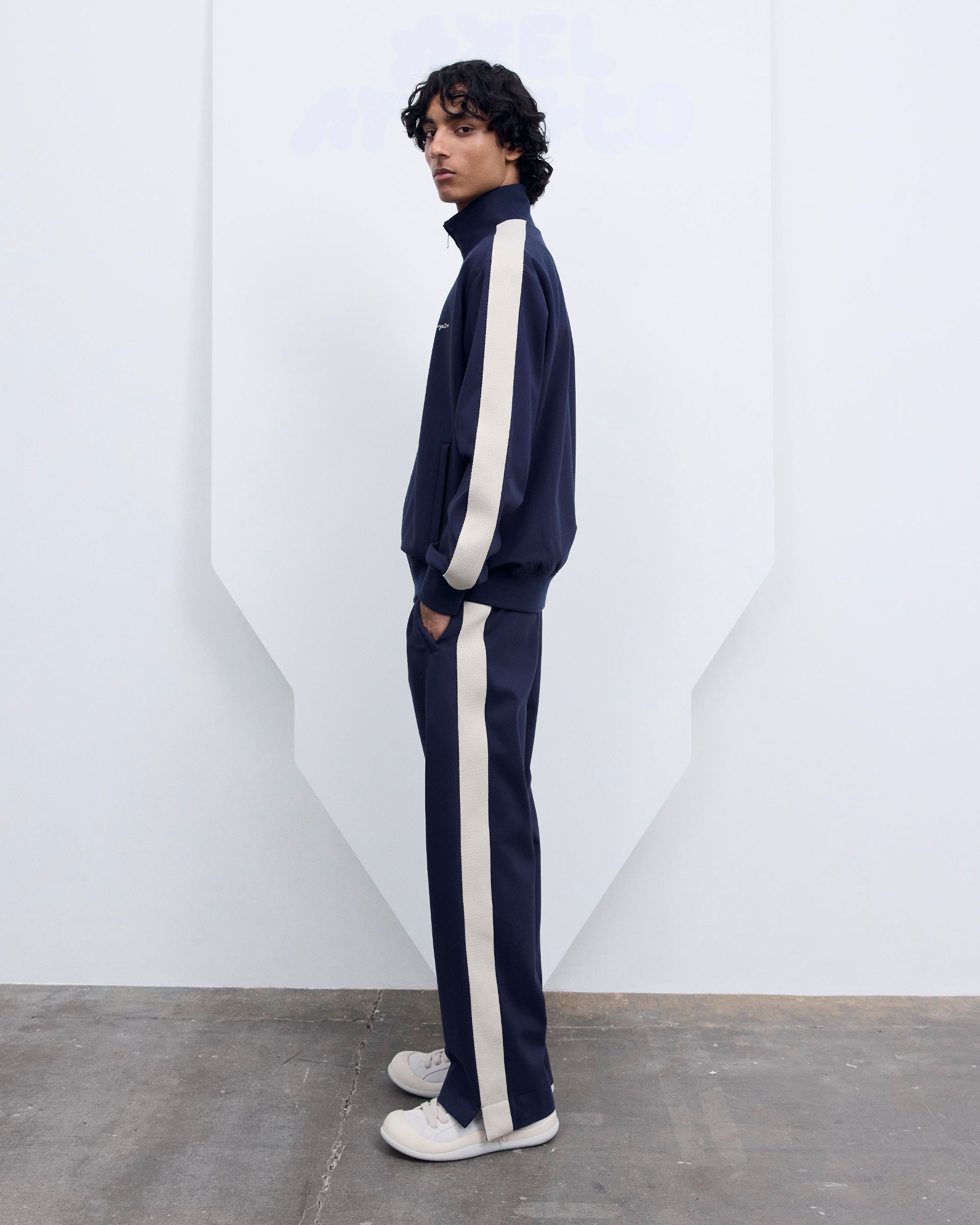 Person wearing a navy tracksuit with white stripes, white sneakers, standing sideways against a minimalistic background.