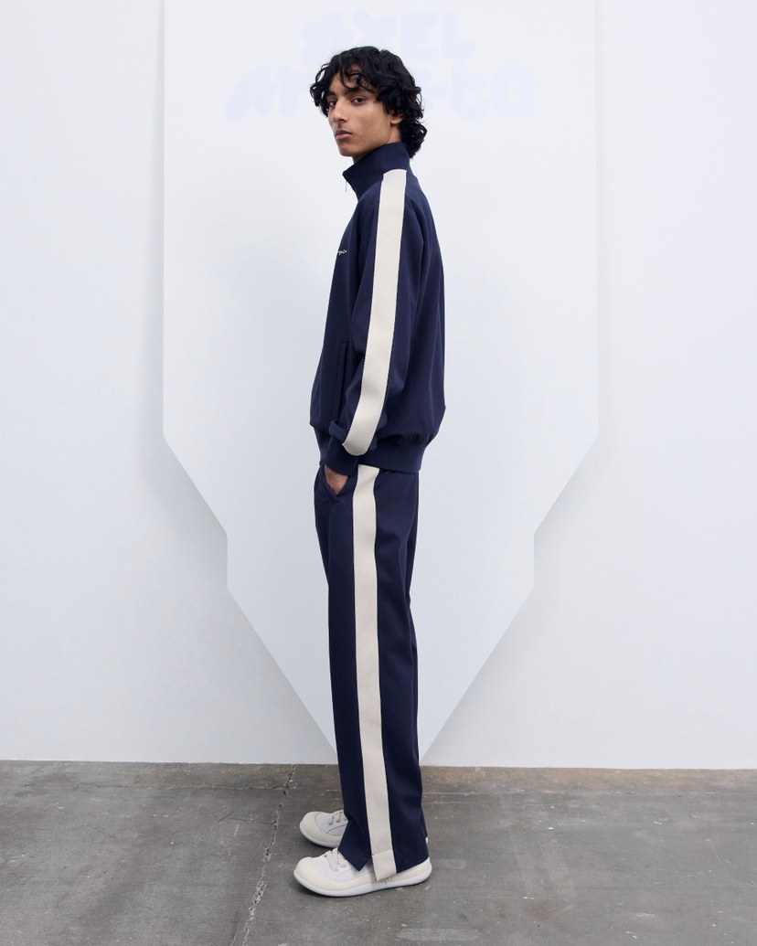 Person wearing a navy tracksuit with white stripes, white sneakers, standing sideways against a minimalistic background.