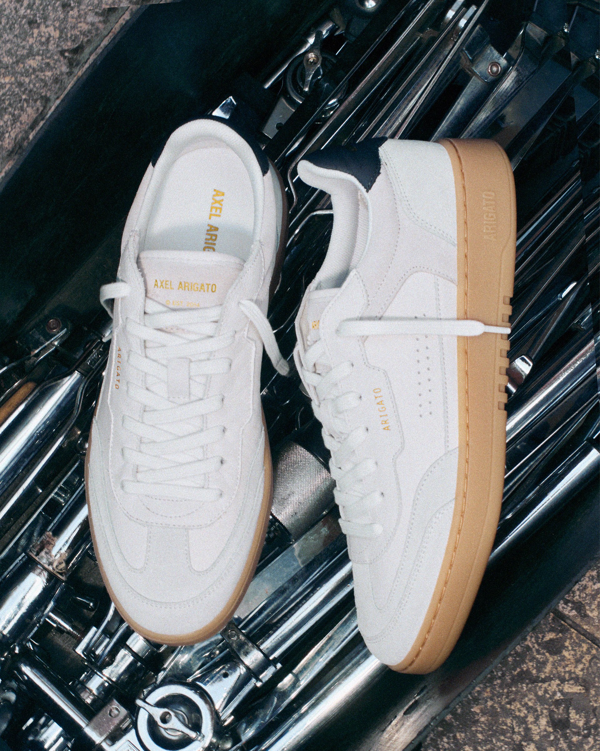 White Axel Arigato sneakers with beige soles, placed on shiny chrome metal, showcasing their sleek design and branding.
