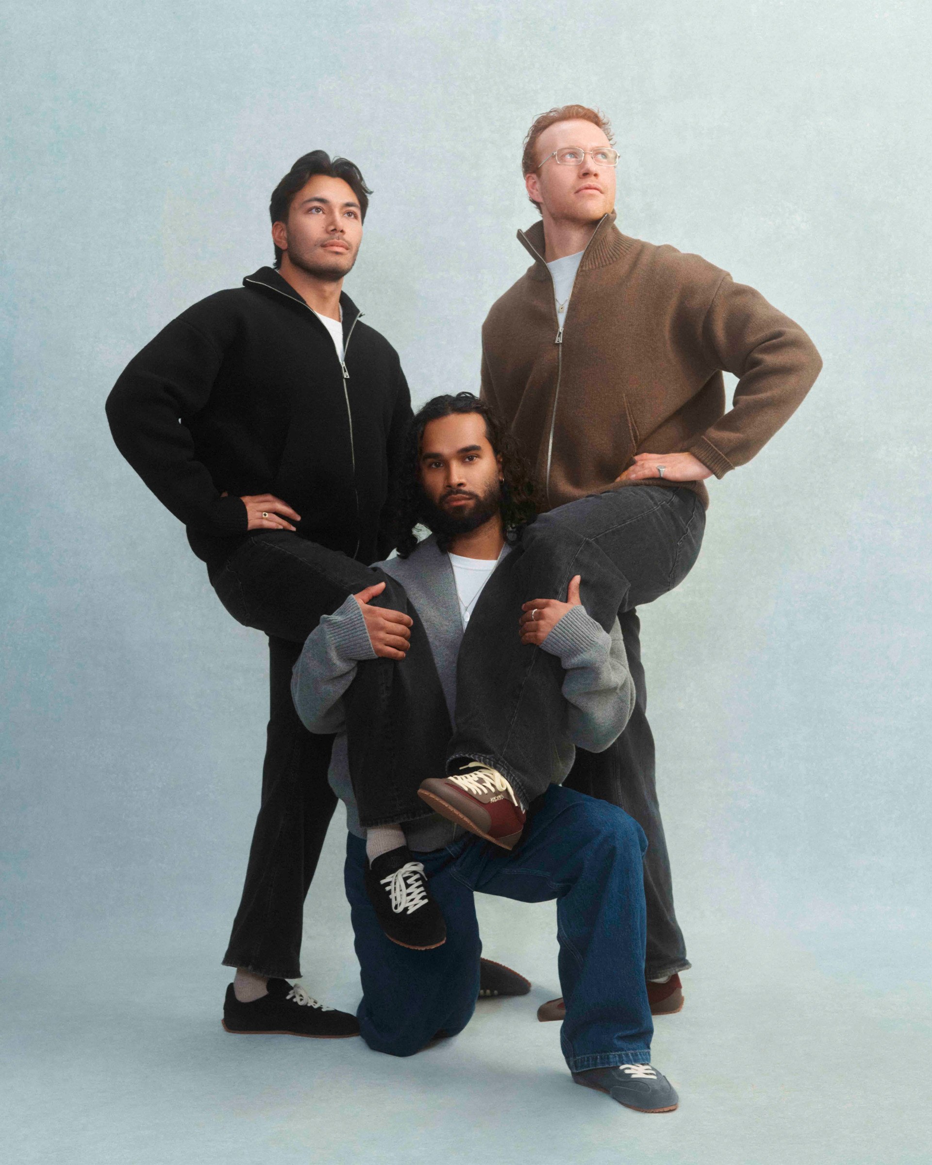Three people pose creatively; one kneels with crossed legs, supporting the feet of two standing individuals against a plain background.