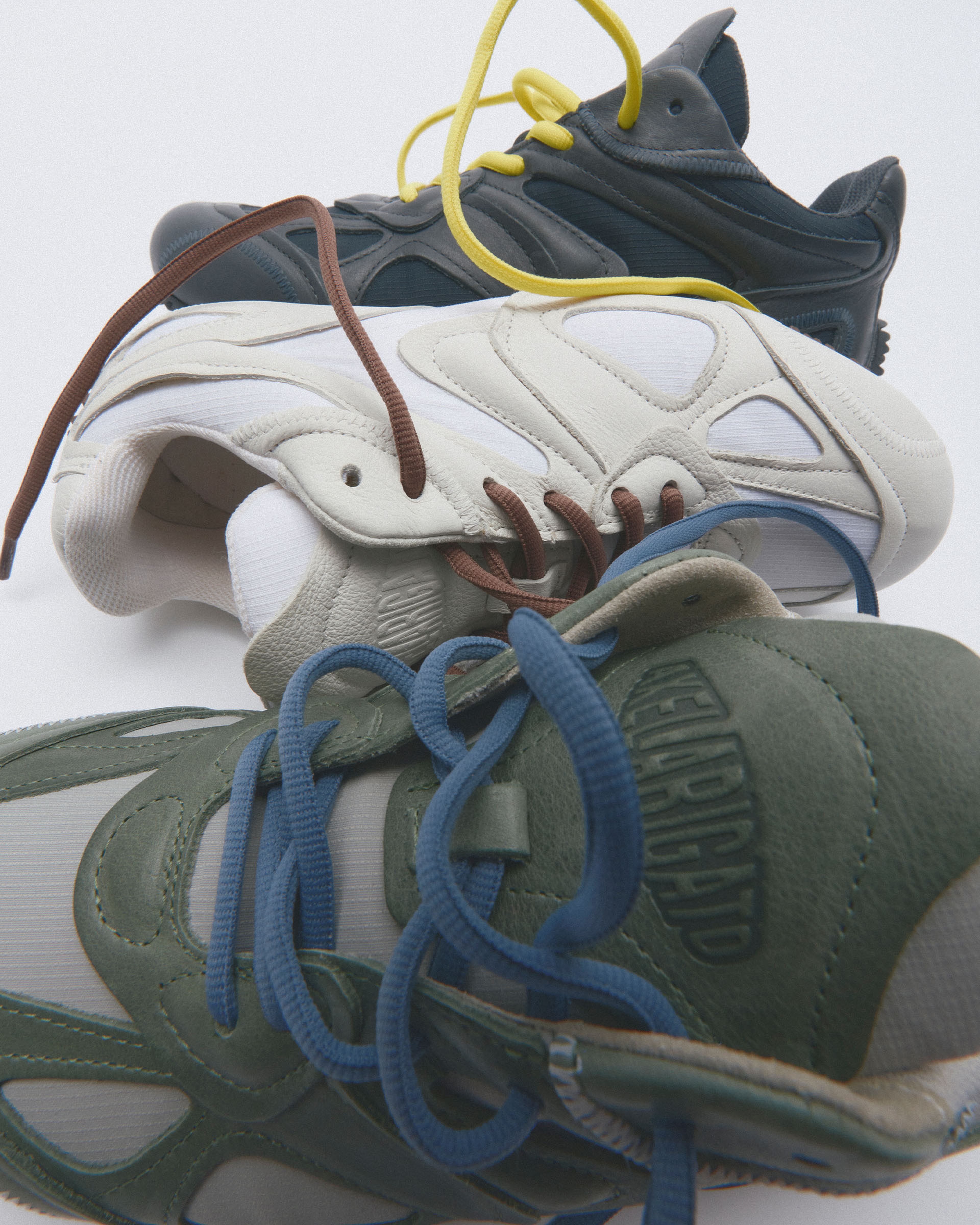 Three stylish sneakers in blue, white, and green colors are intertwined, each with contrasting laces, displayed against a plain background.
