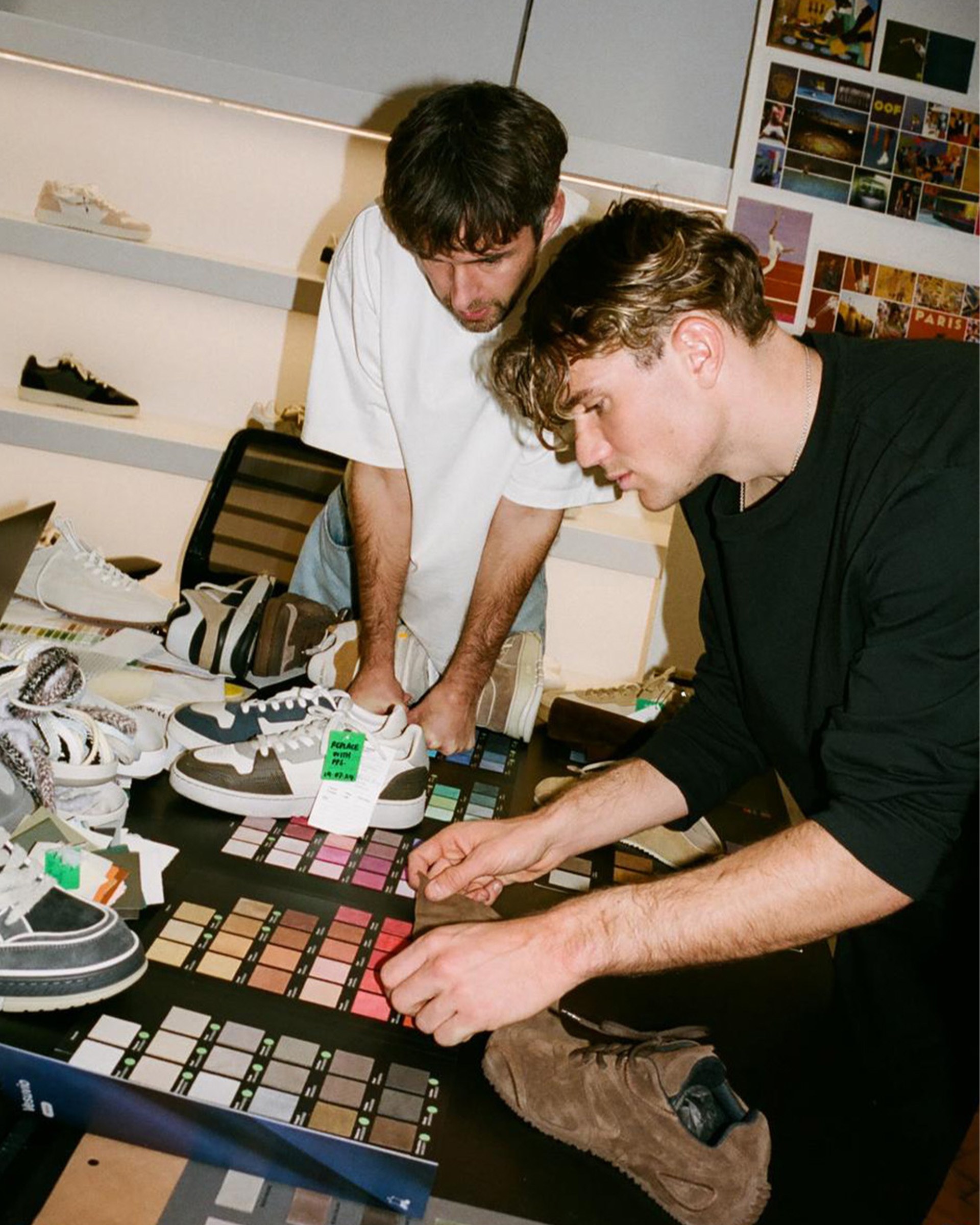 Two men examining color swatches and sneakers on a table in a design studio, with shelves of shoes in the background.