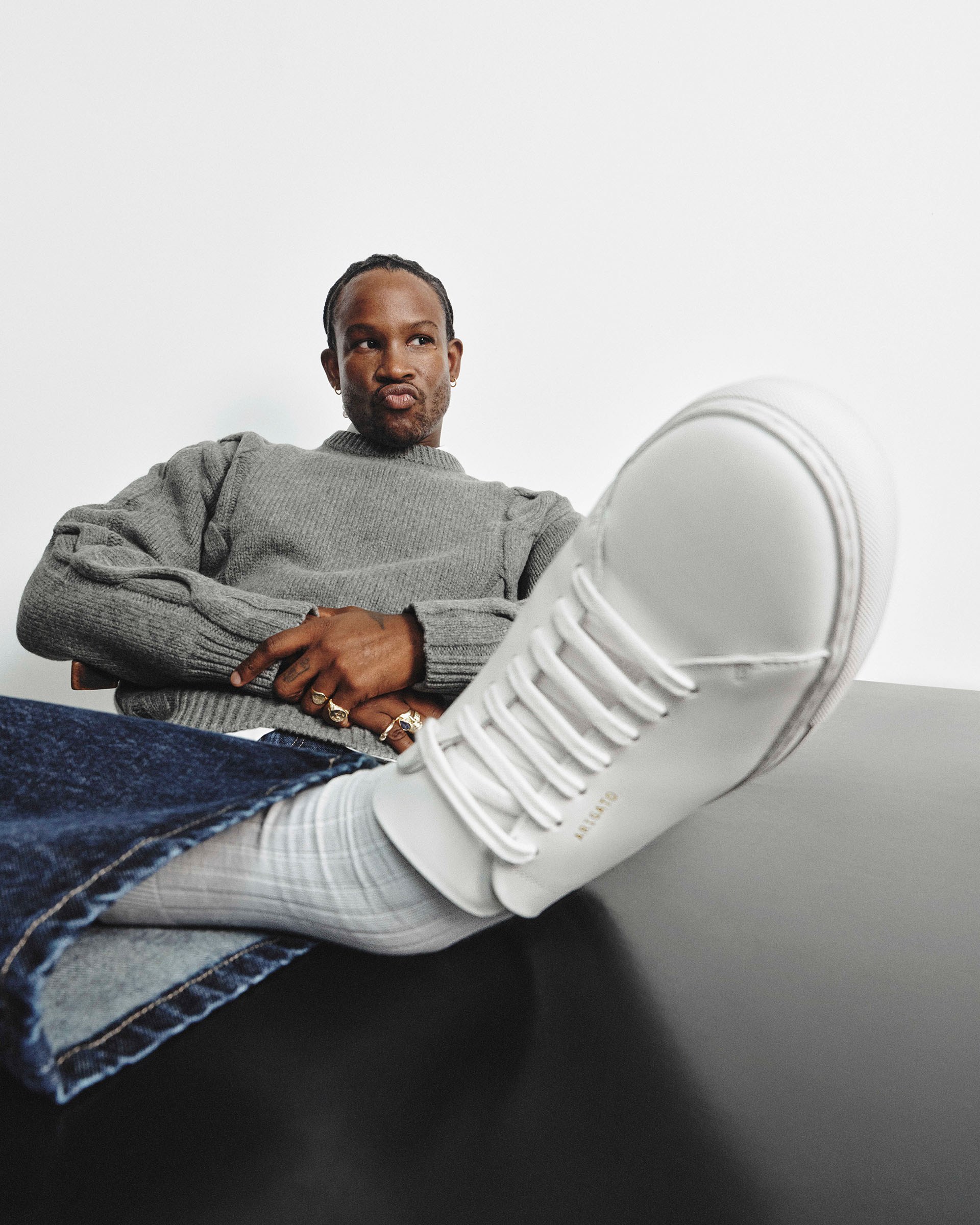Person sitting with a relaxed pose, wearing a gray sweater, jeans, and white sneakers, with one shoe prominently in the foreground.