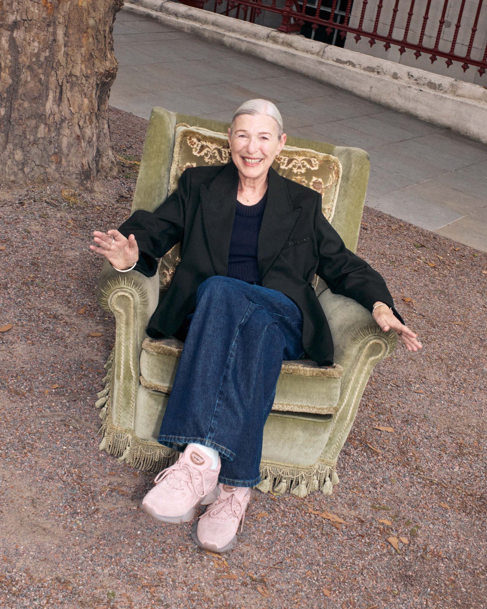 Elderly woman with short gray hair sits smiling in a vintage armchair outdoors, wearing a black jacket, jeans, and pink sneakers.