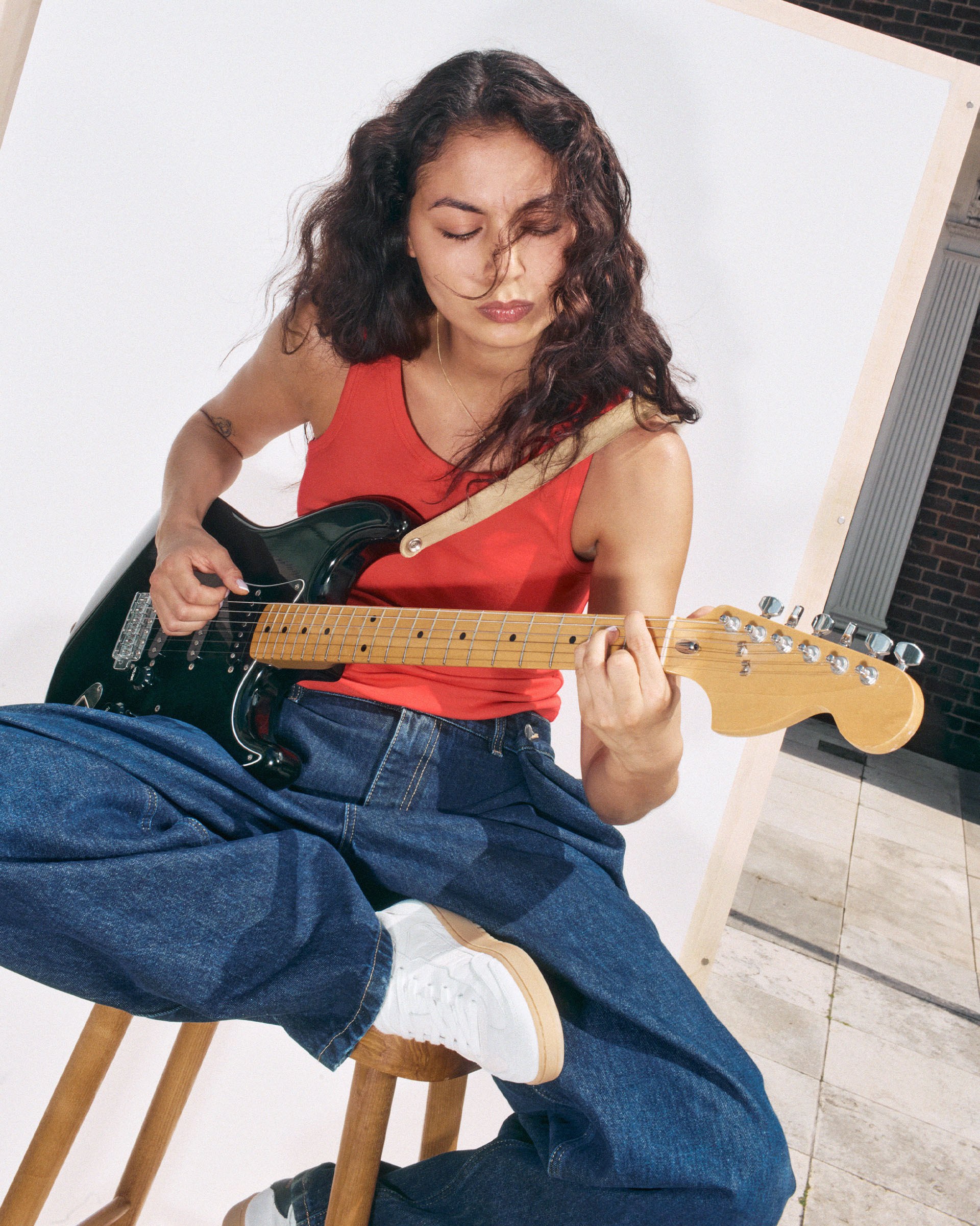Person with long hair playing an electric guitar, wearing a red tank top and jeans, seated on a stool against a neutral background.