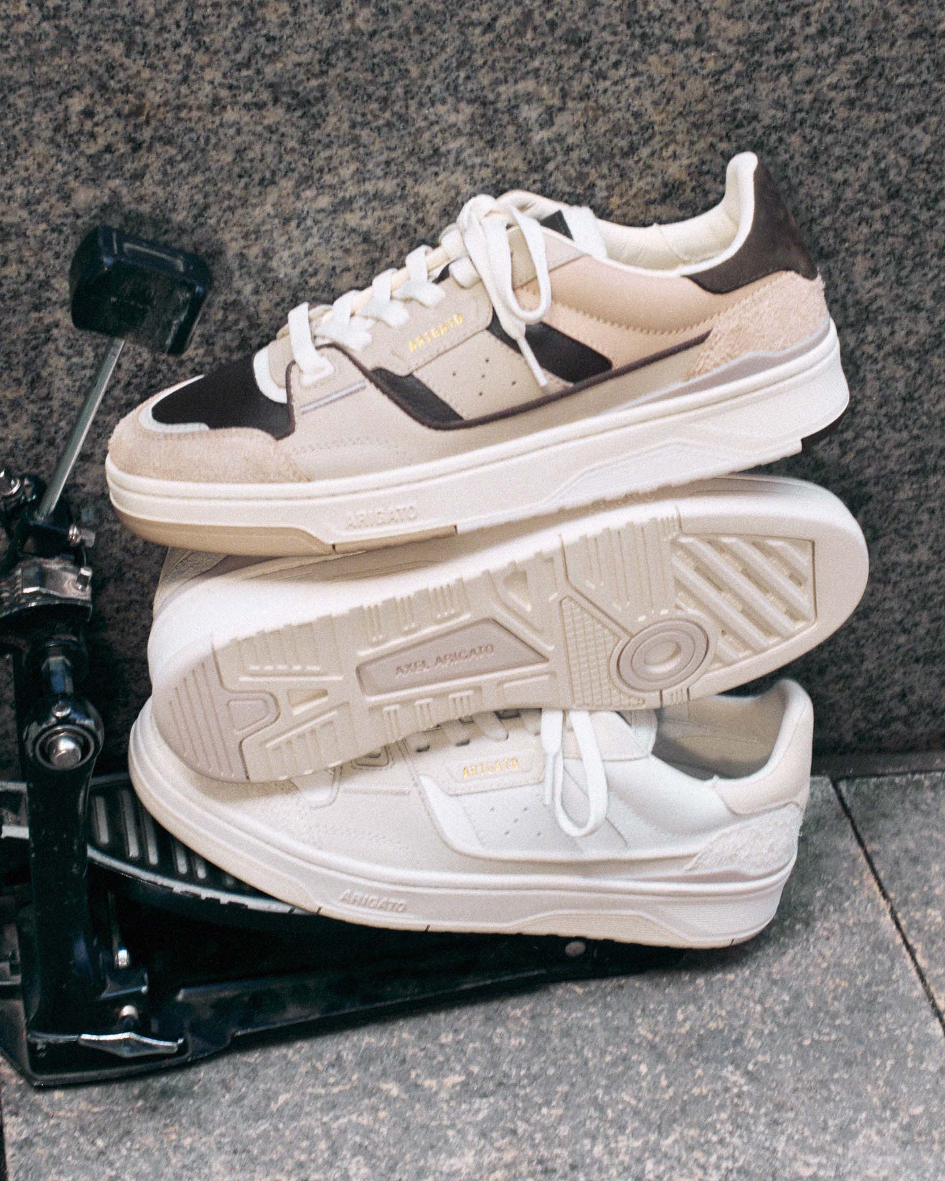 Two stacked sneakers on a metal stand; one in brown and beige, the other in white, with textured soles and "Axel Arigato" branding.