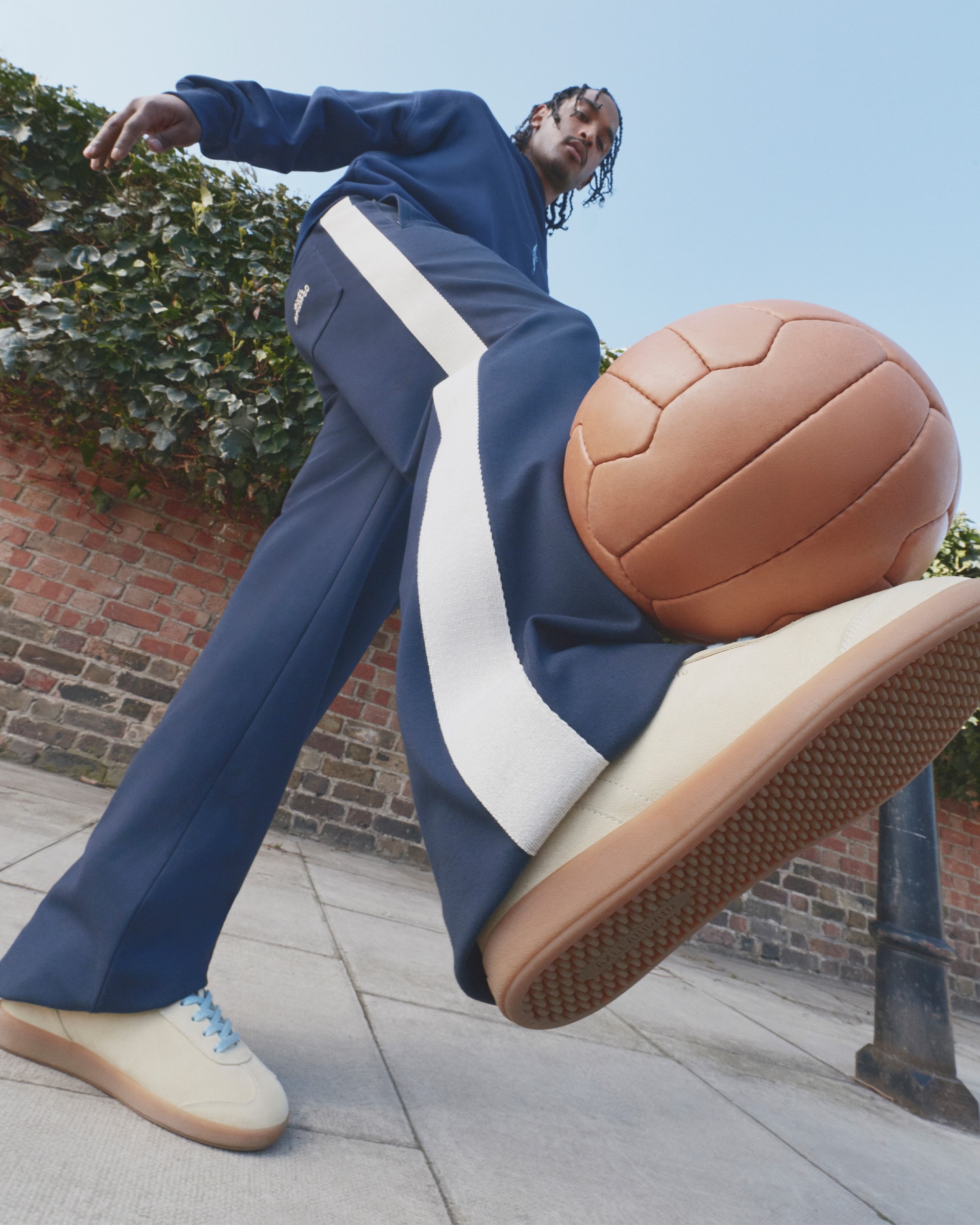 Person in a blue tracksuit kicks a large brown soccer ball outdoors, with a brick wall and ivy in the background. Low-angle view.