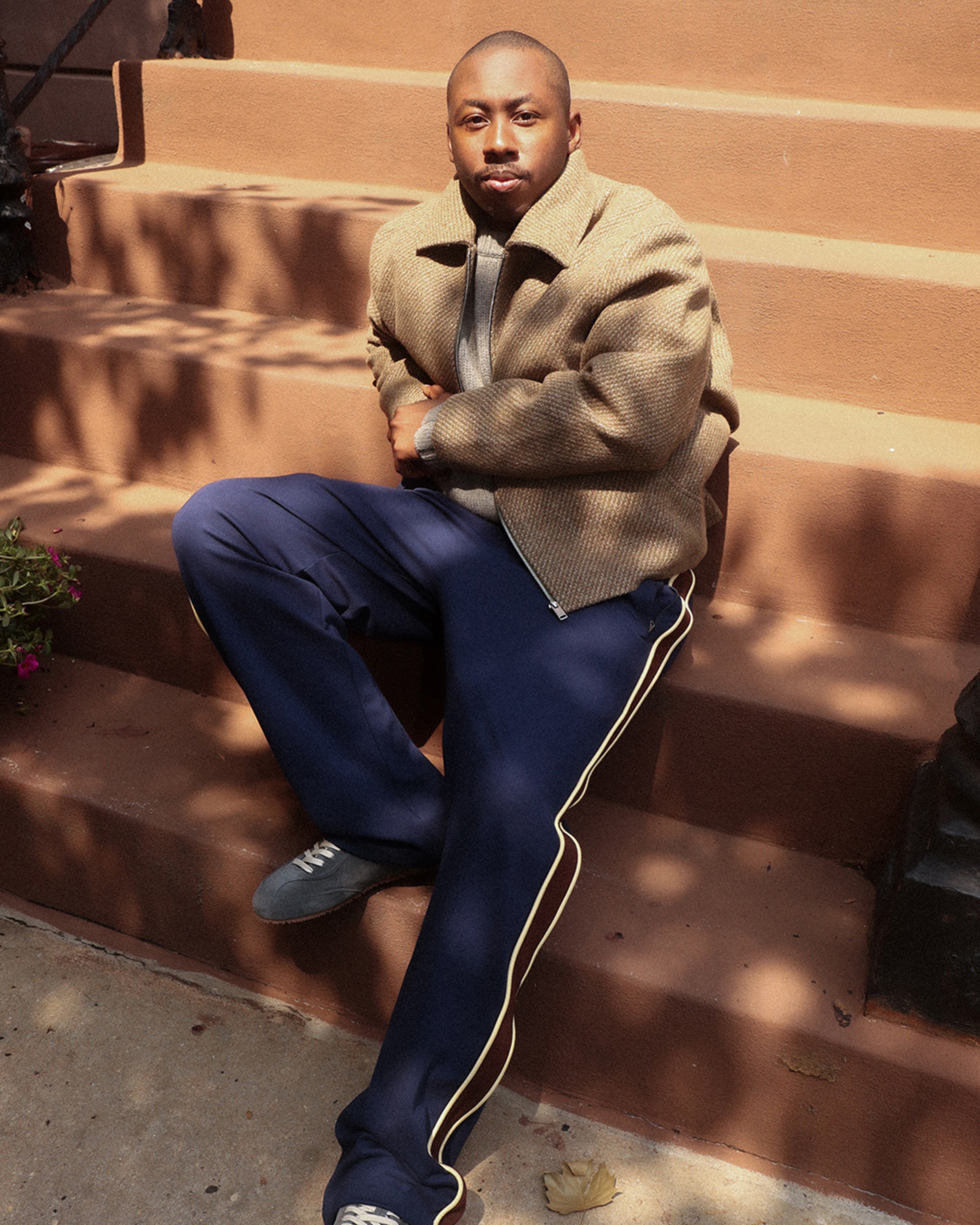 A person sits on outdoor steps wearing a brown jacket, gray shirt, and blue pants with a white stripe, with sunlight casting shadows around them.