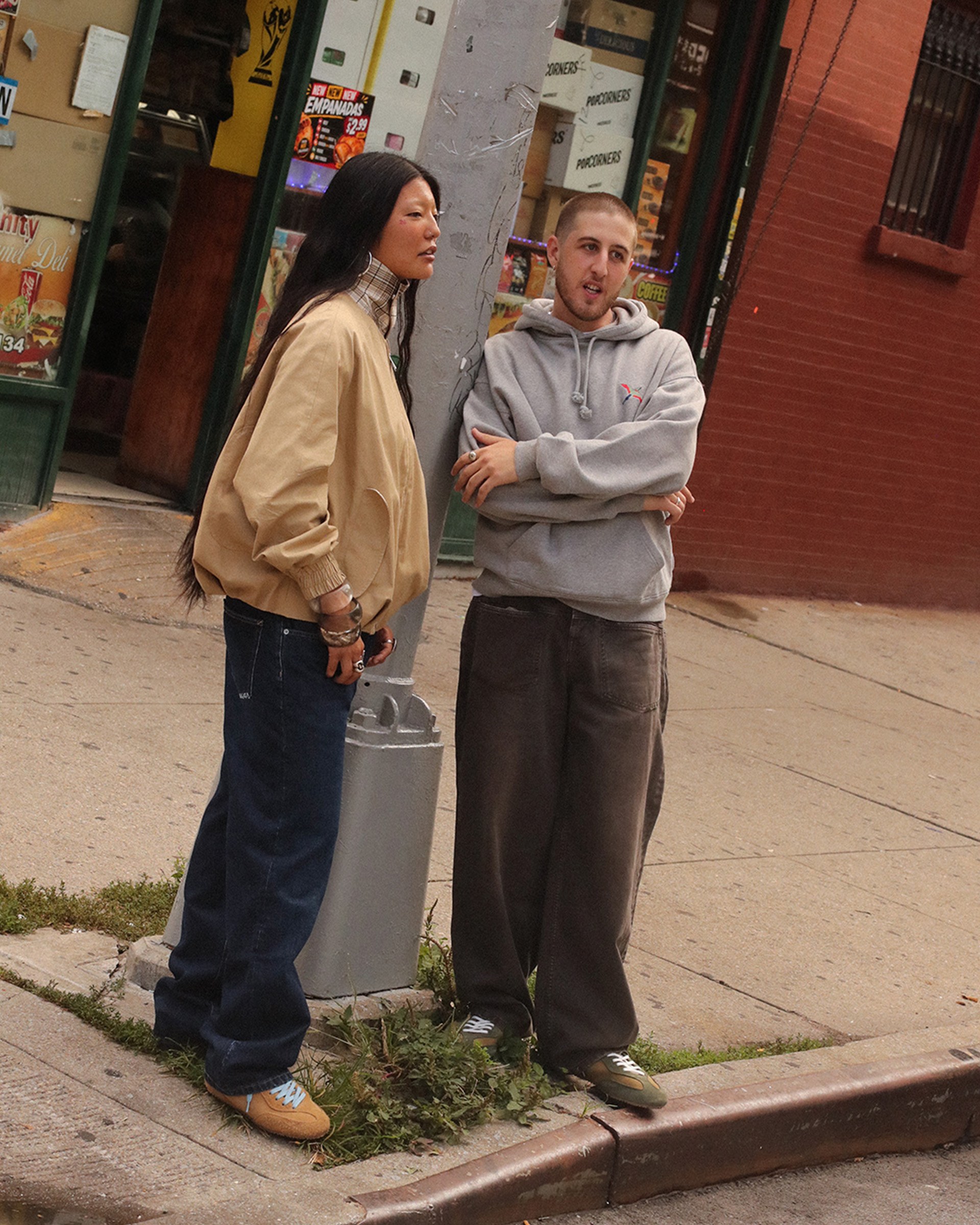 Two people stand on a street corner, one in a tan jacket and jeans, the other in a gray hoodie and baggy pants, in an urban setting.