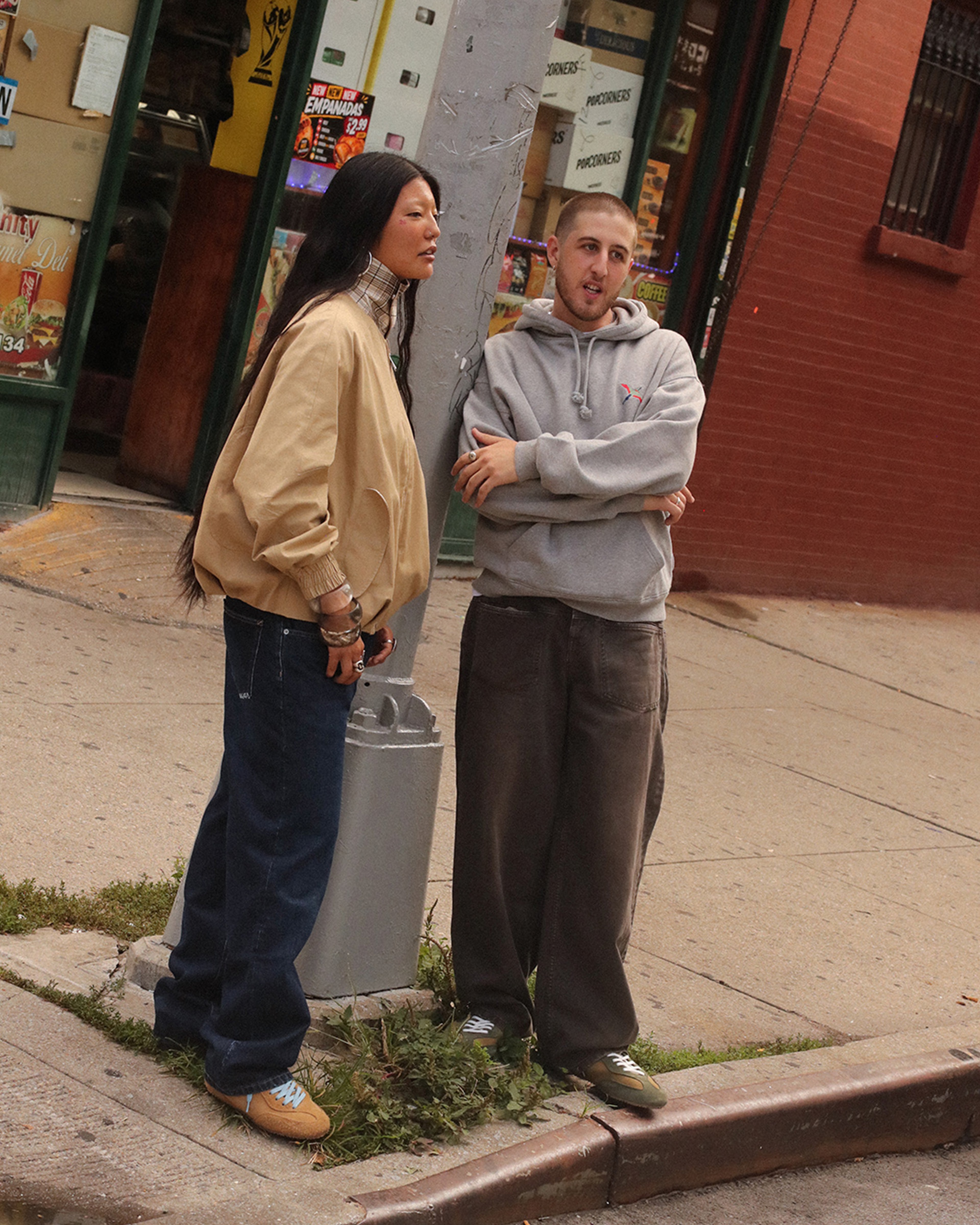 Two people stand on a street corner, one in a tan jacket and jeans, the other in a gray hoodie and baggy pants, in an urban setting.