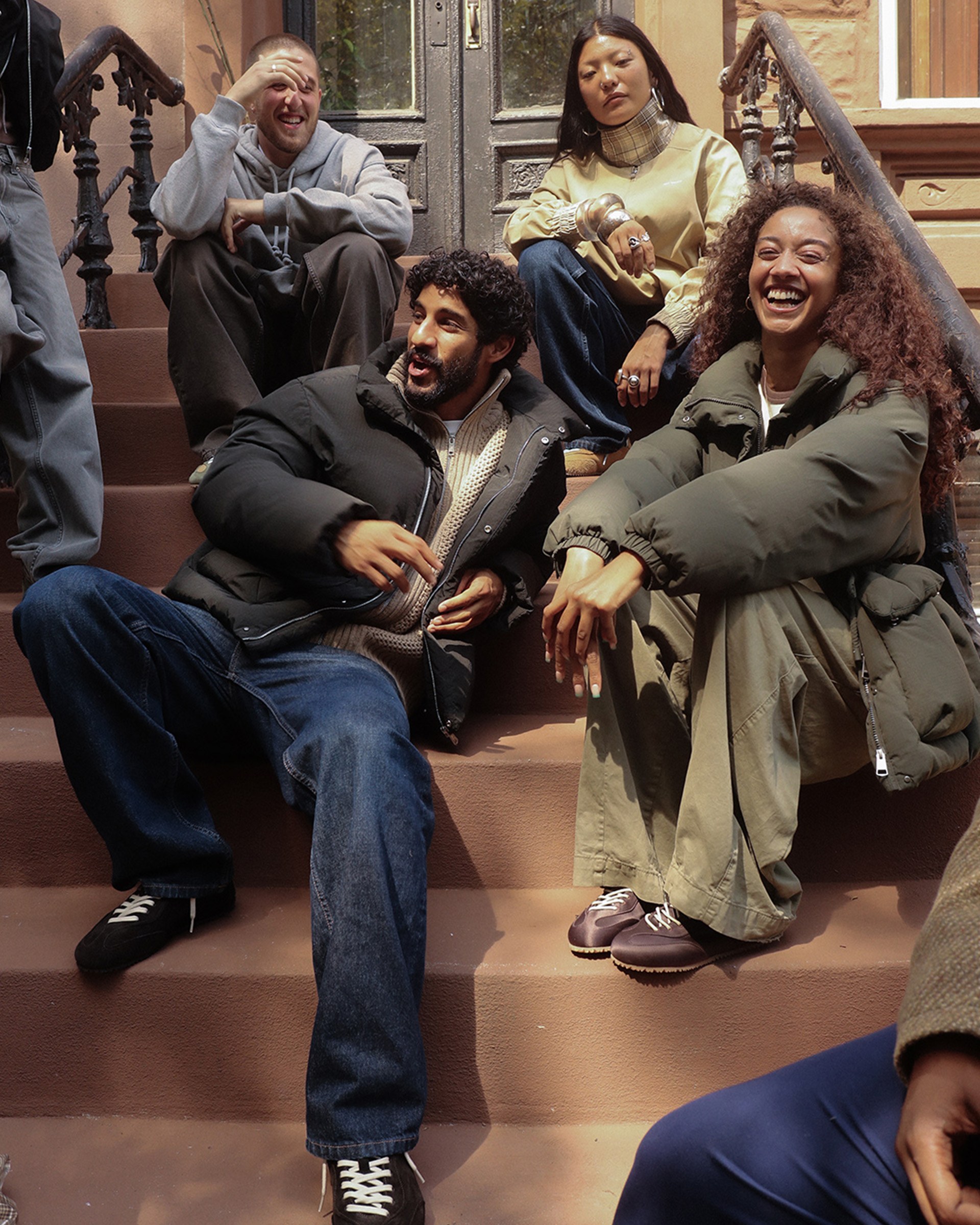 Four people sitting casually and smiling on outdoor steps, dressed warmly, one laughing and gesturing, with a brick building in the background.