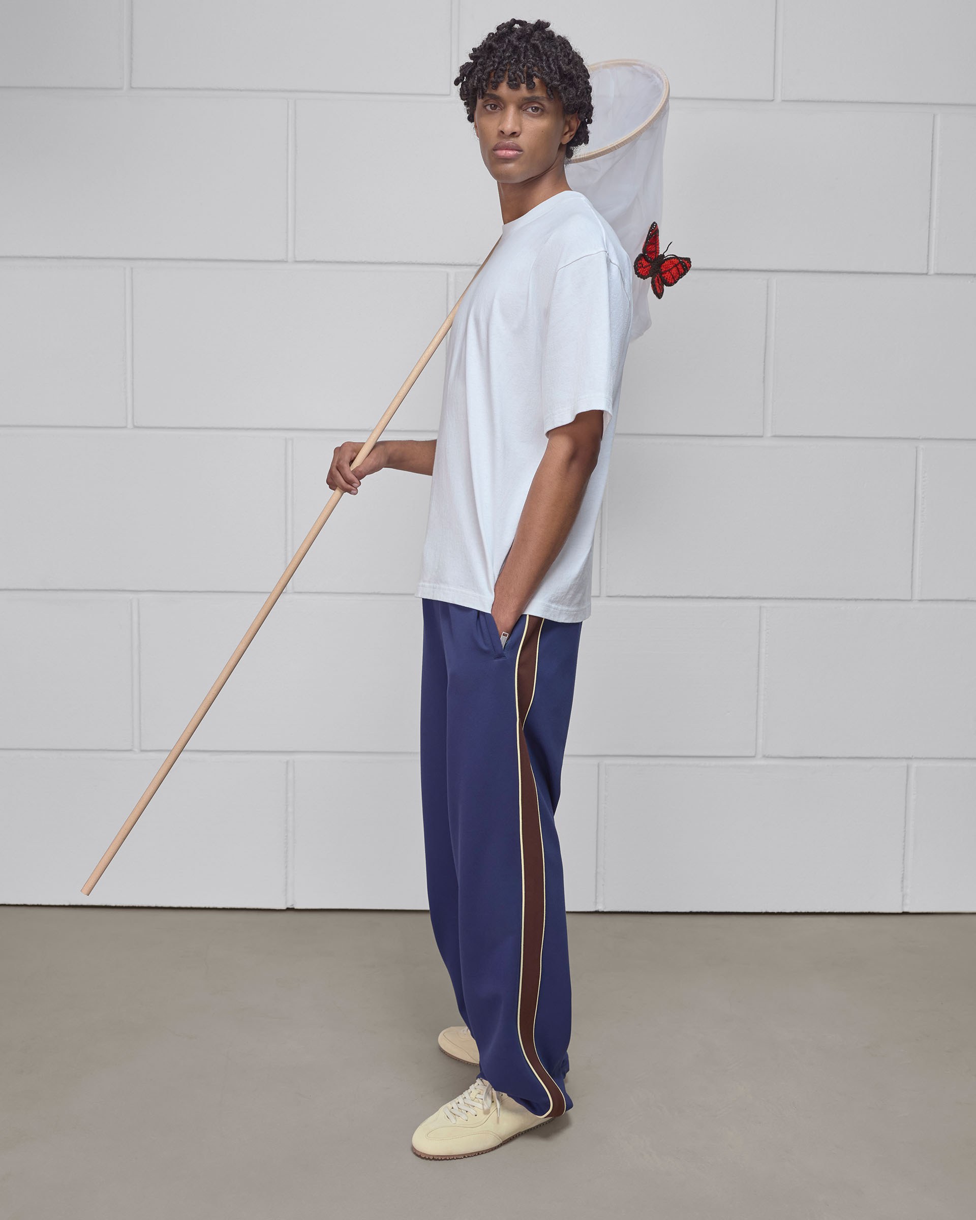 Man holding a butterfly net with a red butterfly on it, wearing a white t-shirt and blue track pants, standing against a tiled wall.