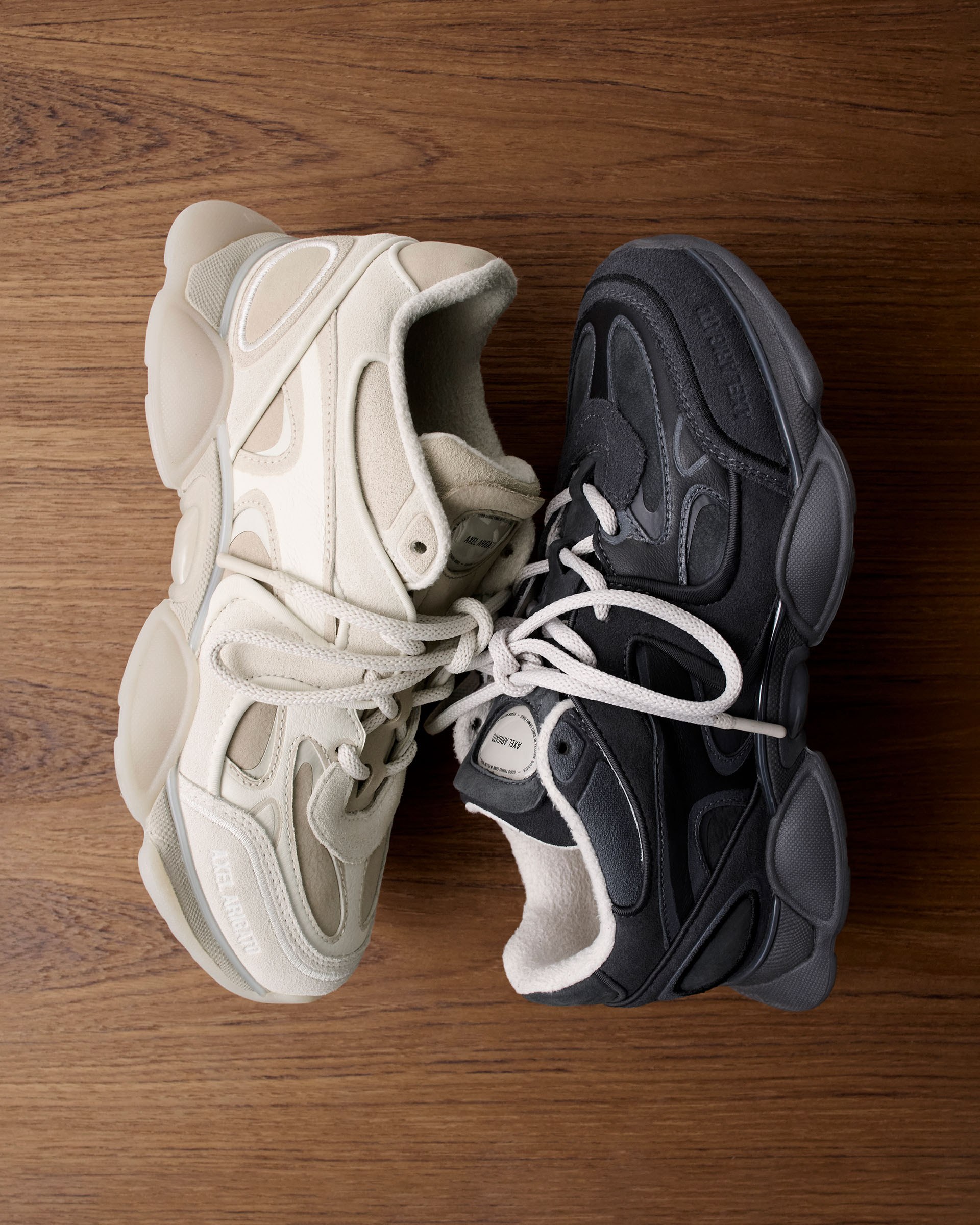 Two sneakers on a wooden surface, one cream-colored and the other dark gray, with chunky soles and intricate panel designs.