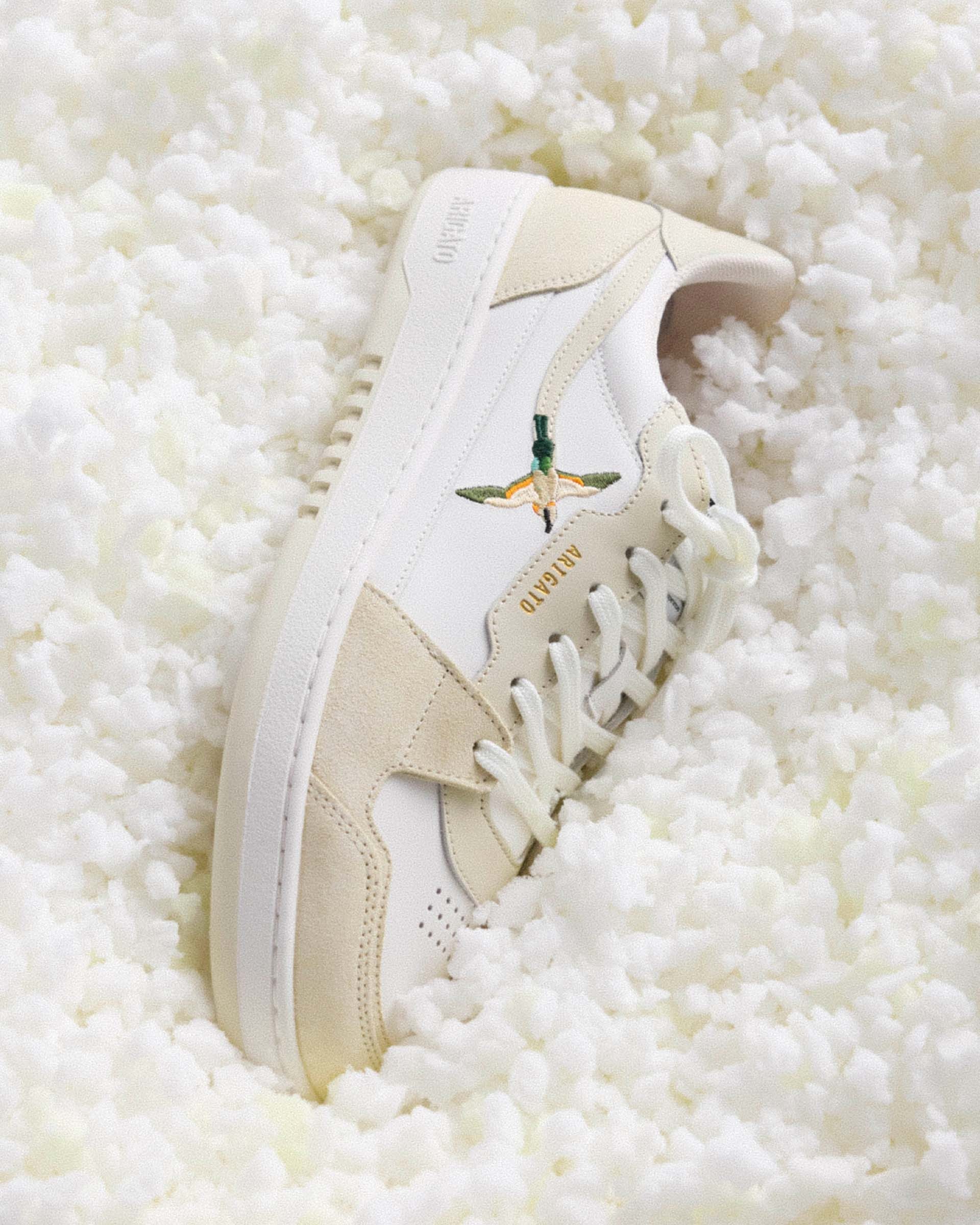 White and beige sneaker with a bird logo on the side, placed on a textured white surface.
