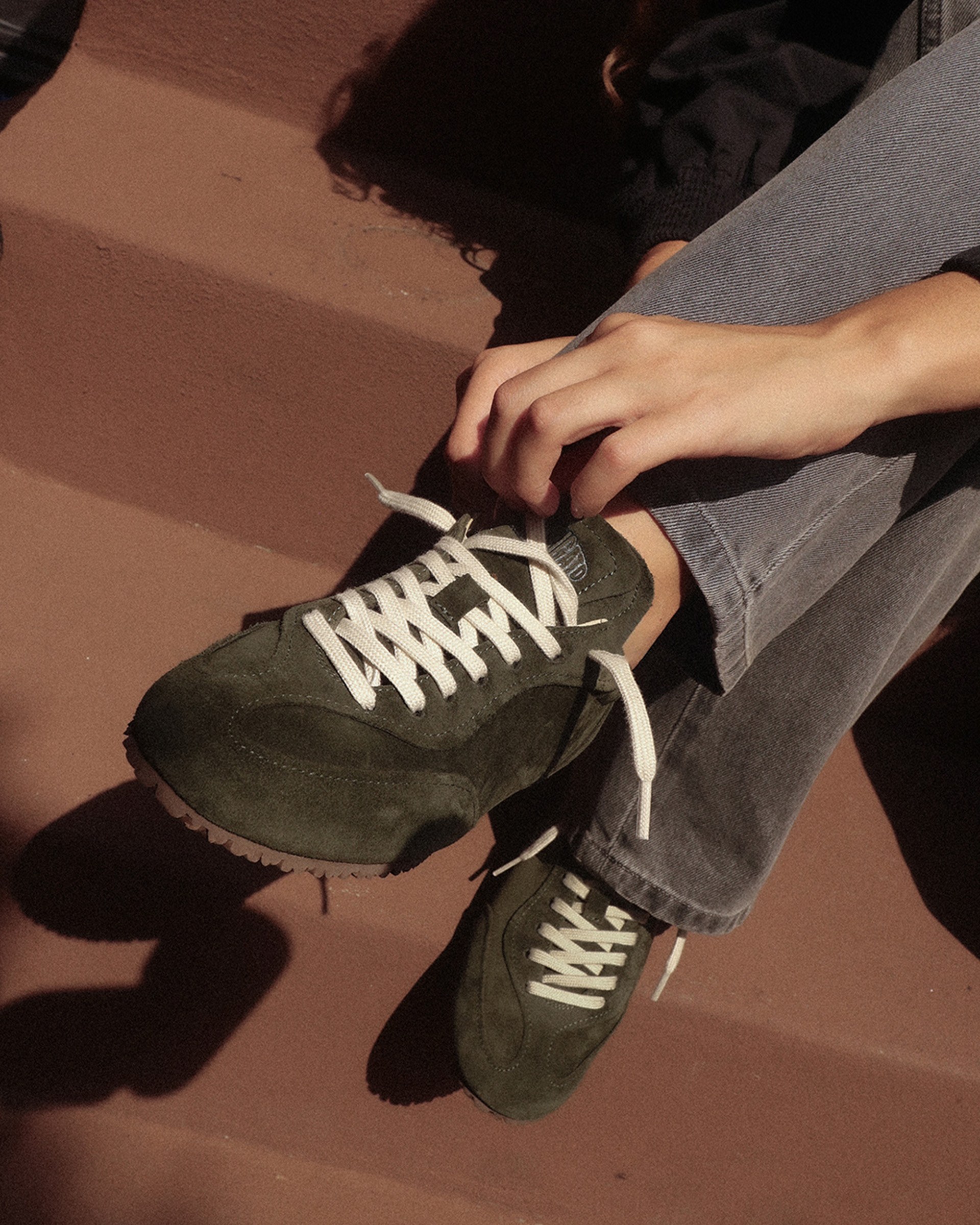 Person sitting on steps, wearing green suede sneakers with white laces, adjusting shoelaces, and dressed in blue jeans.