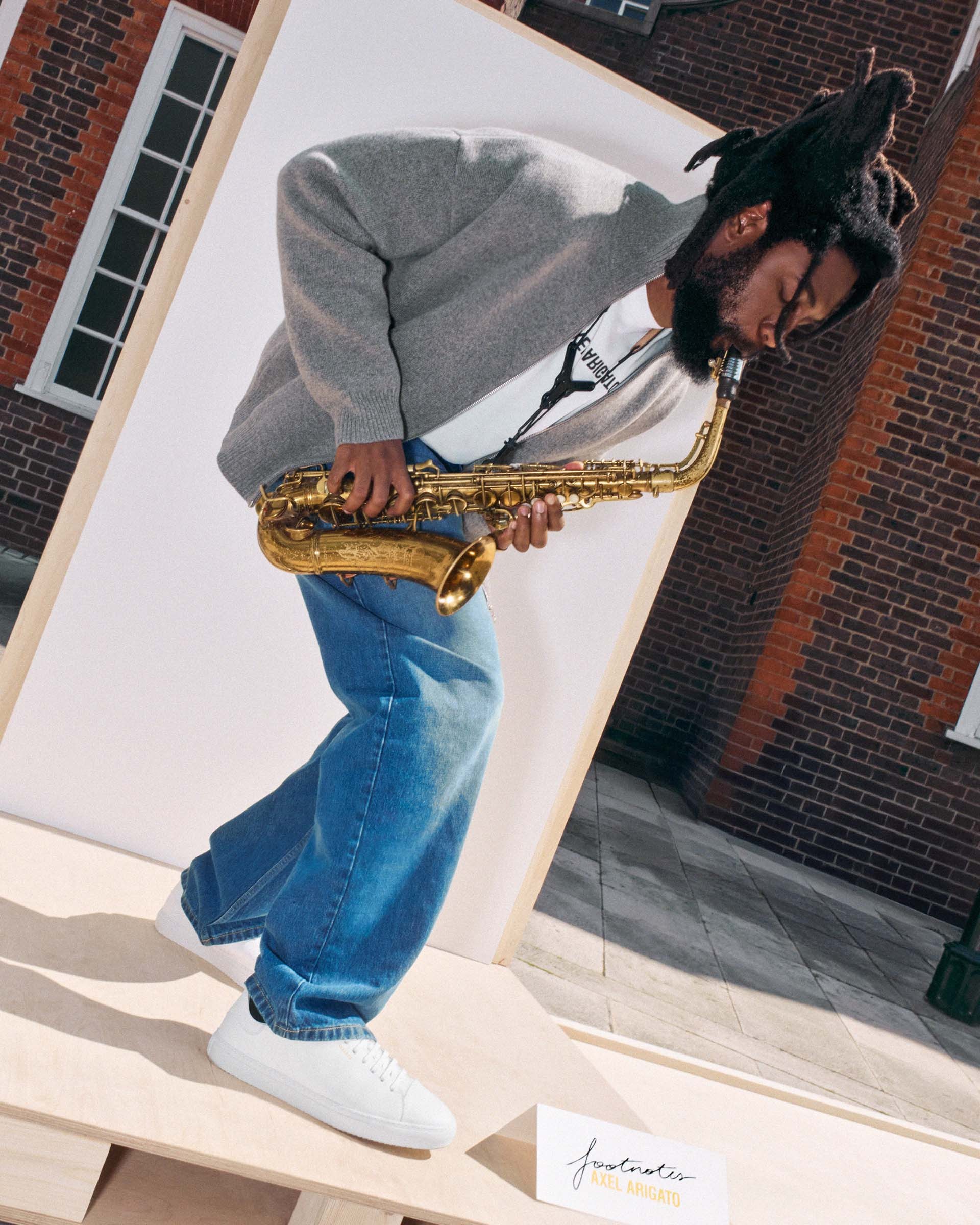 Person with dreadlocks playing a saxophone, wearing a gray sweater, blue jeans, and white sneakers, standing on a wooden platform outdoors.