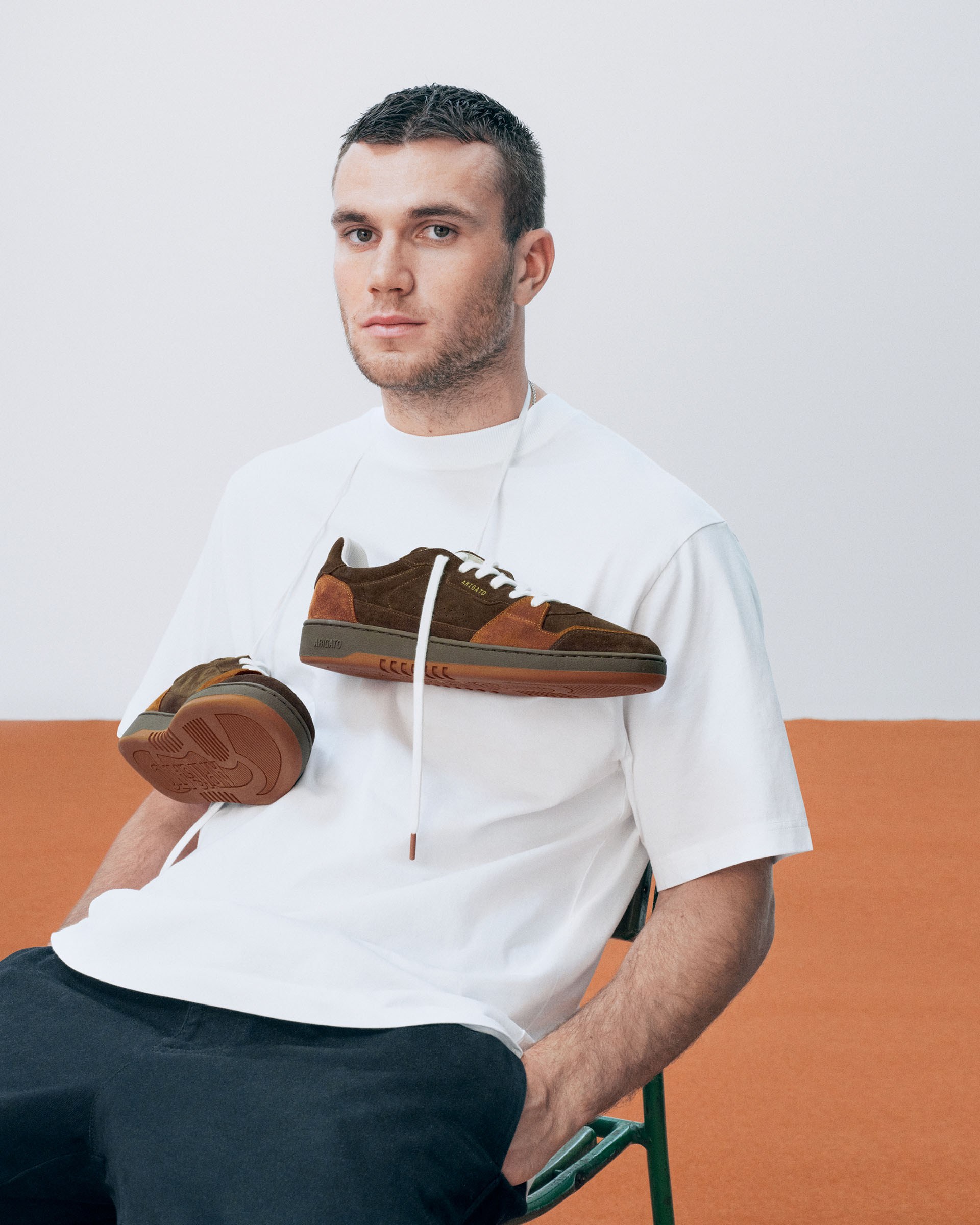 Man in a white T-shirt and dark pants sitting on a chair with brown sneakers hanging from his neck, against a minimalist background.