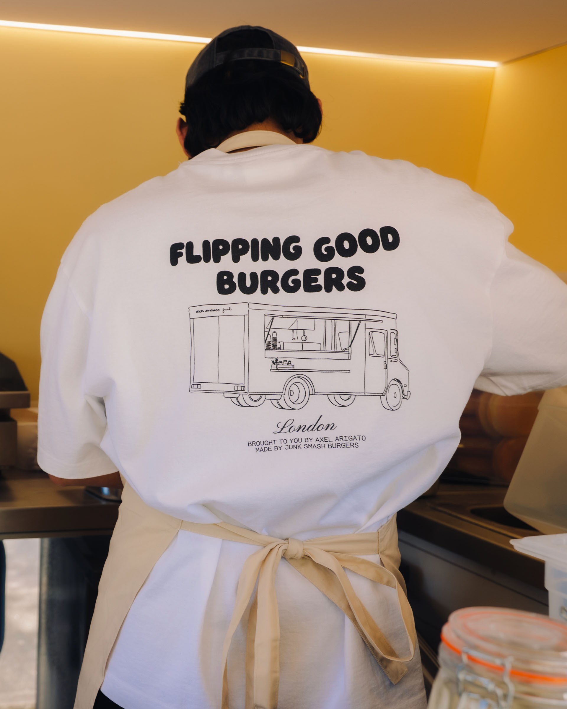 Person in a white "Flipping Good Burgers" shirt, working at a food truck. The shirt has a simple burger truck illustration.