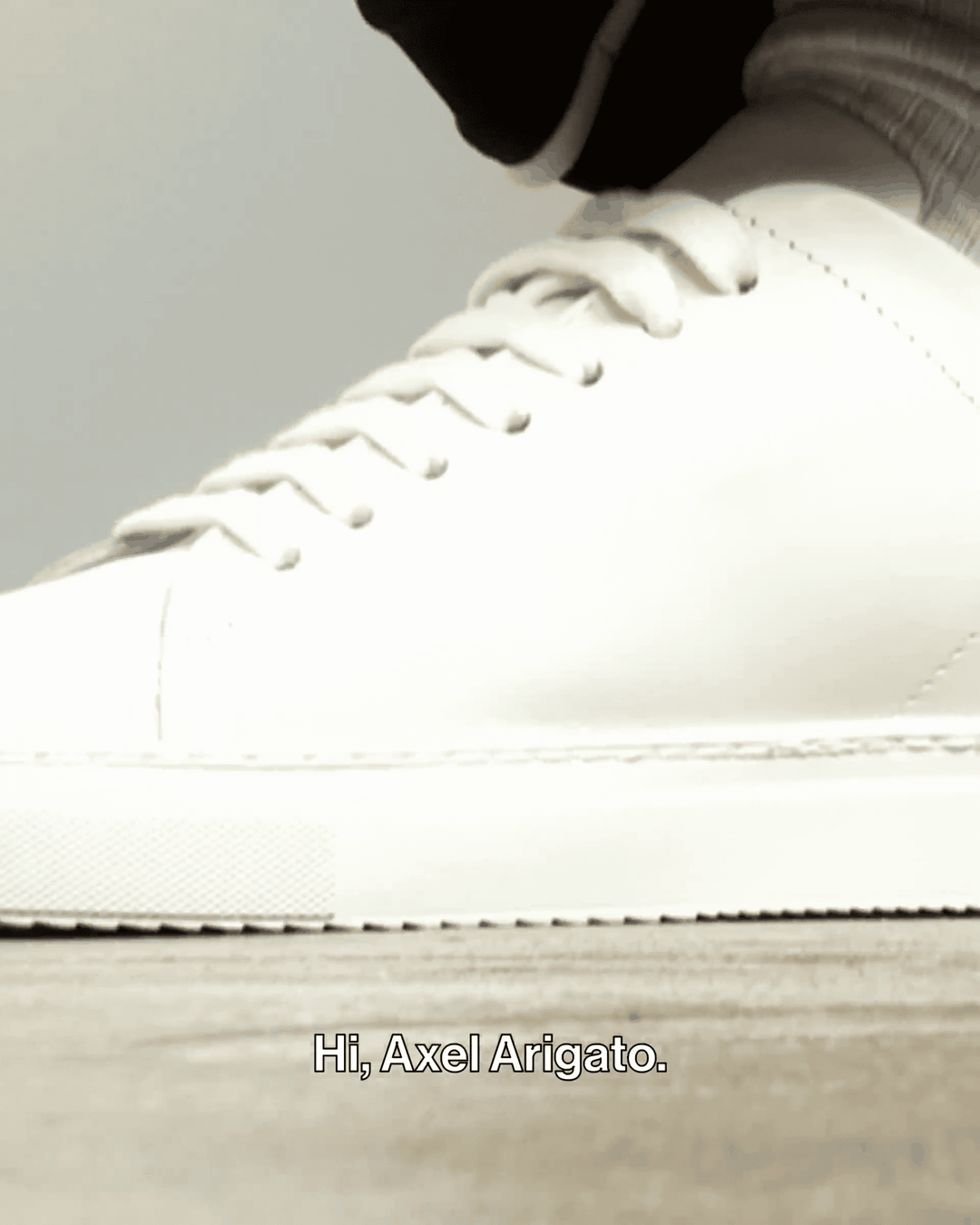 Close-up of a white sneaker with a thick sole on a wooden surface. Text at the bottom reads, "Hi, Axel Arigato."