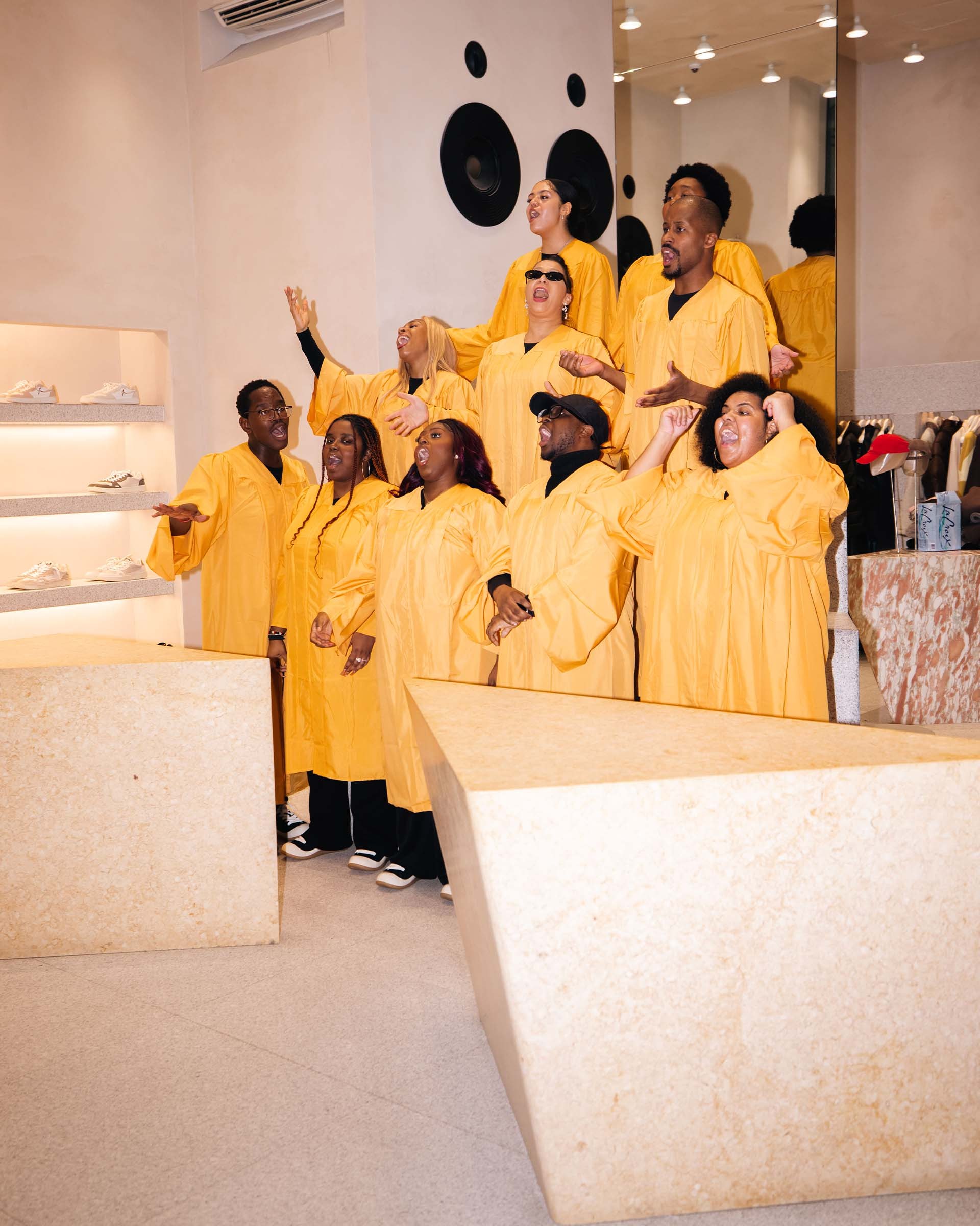 A group of people in yellow robes sing joyfully in a modern room with geometric decor and shoes displayed on shelves.