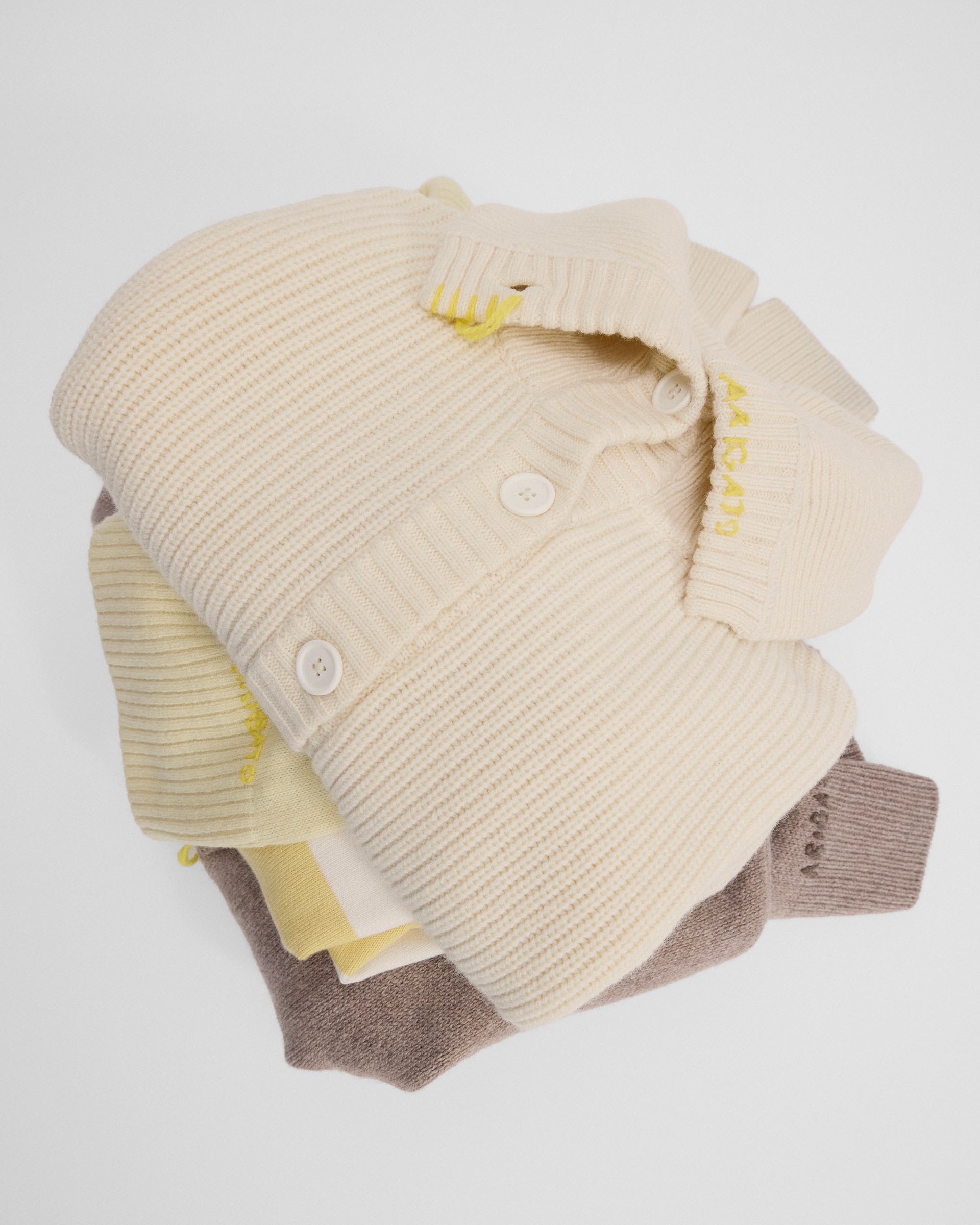A stack of folded sweaters in beige and brown tones, with ribbed textures and buttons, placed on a white background.