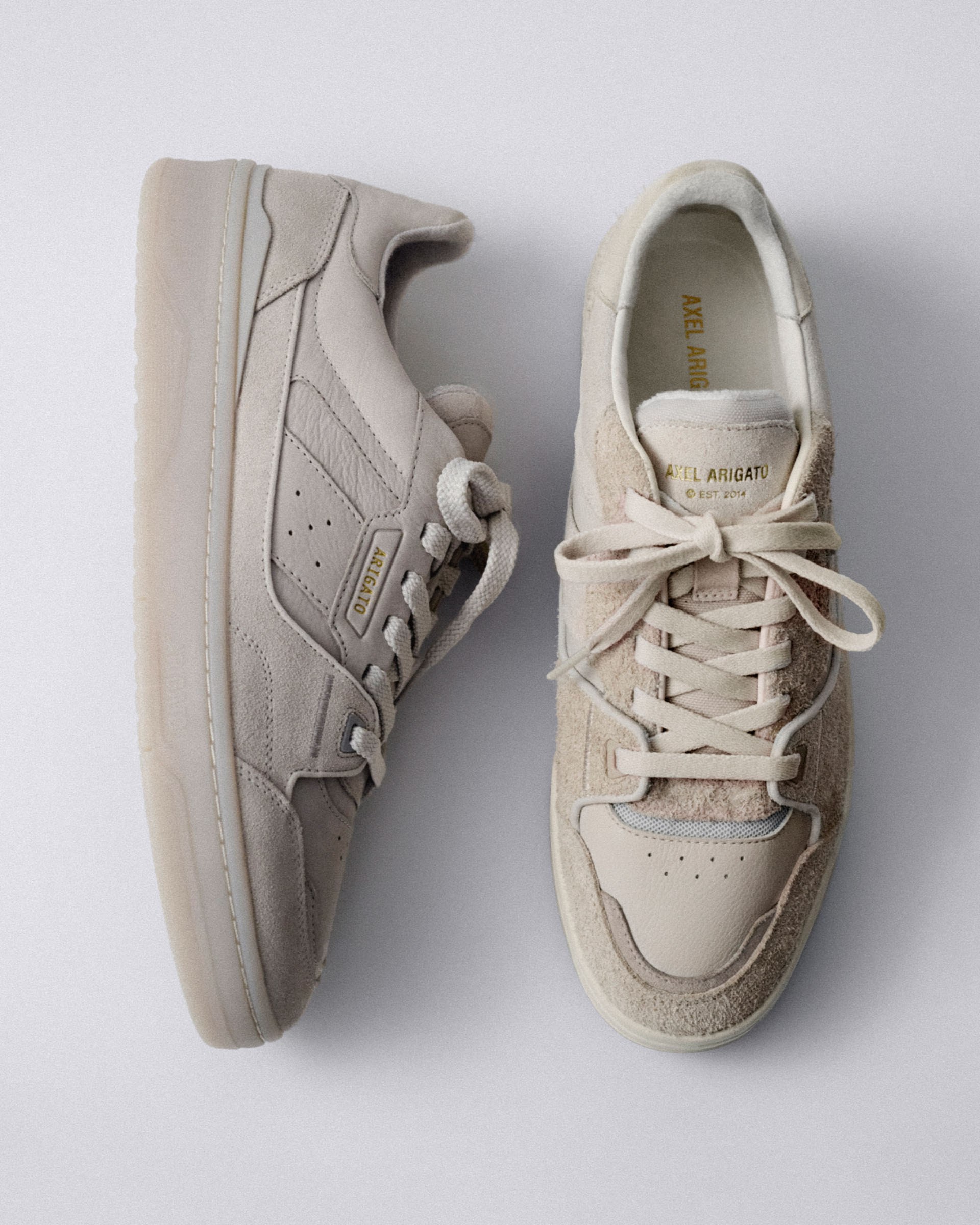 Pair of beige sneakers, one laid sideways and the other top-down, on a light gray background, showcasing suede and leather textures.
