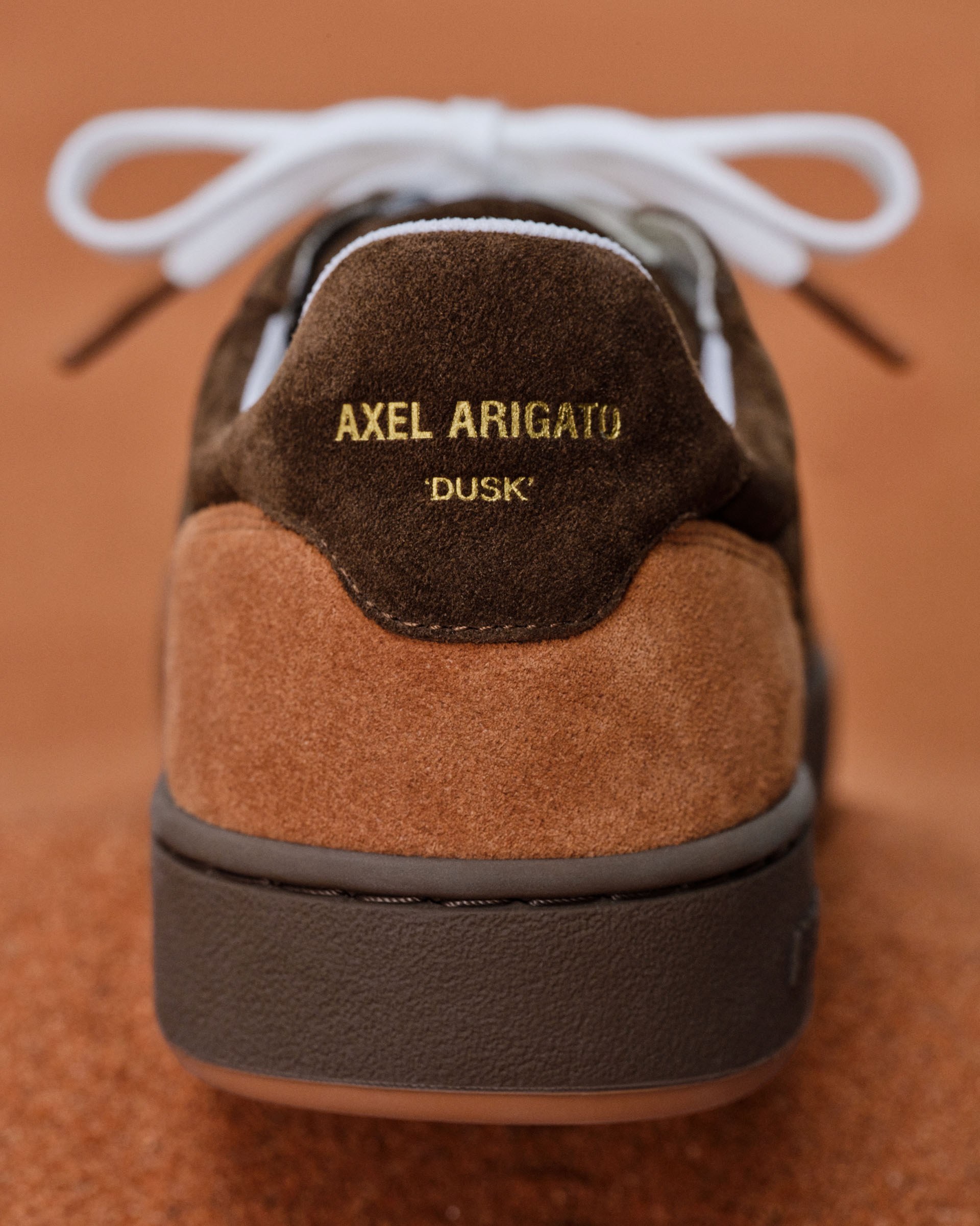 Close-up of a brown suede sneaker with white laces, featuring the gold text "AXEL ARIGATO 'DUSK'" on the heel against a blurred background.