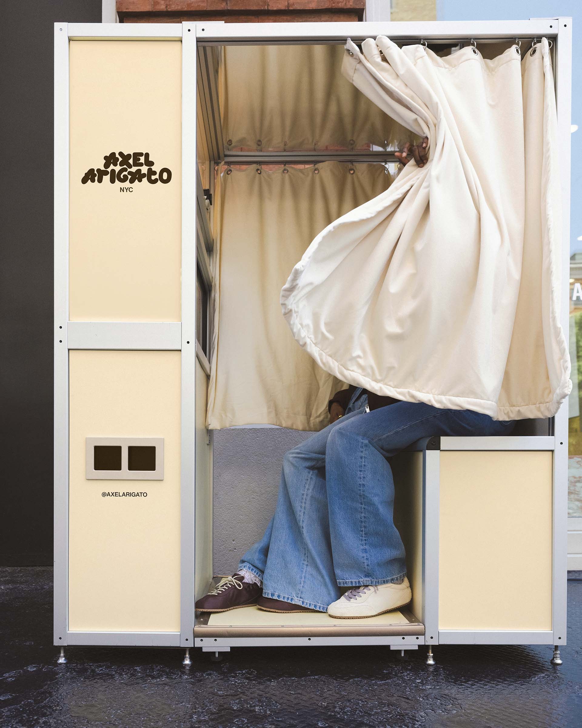 A person wearing denim jeans and sneakers sits inside a photo booth with cream curtains.