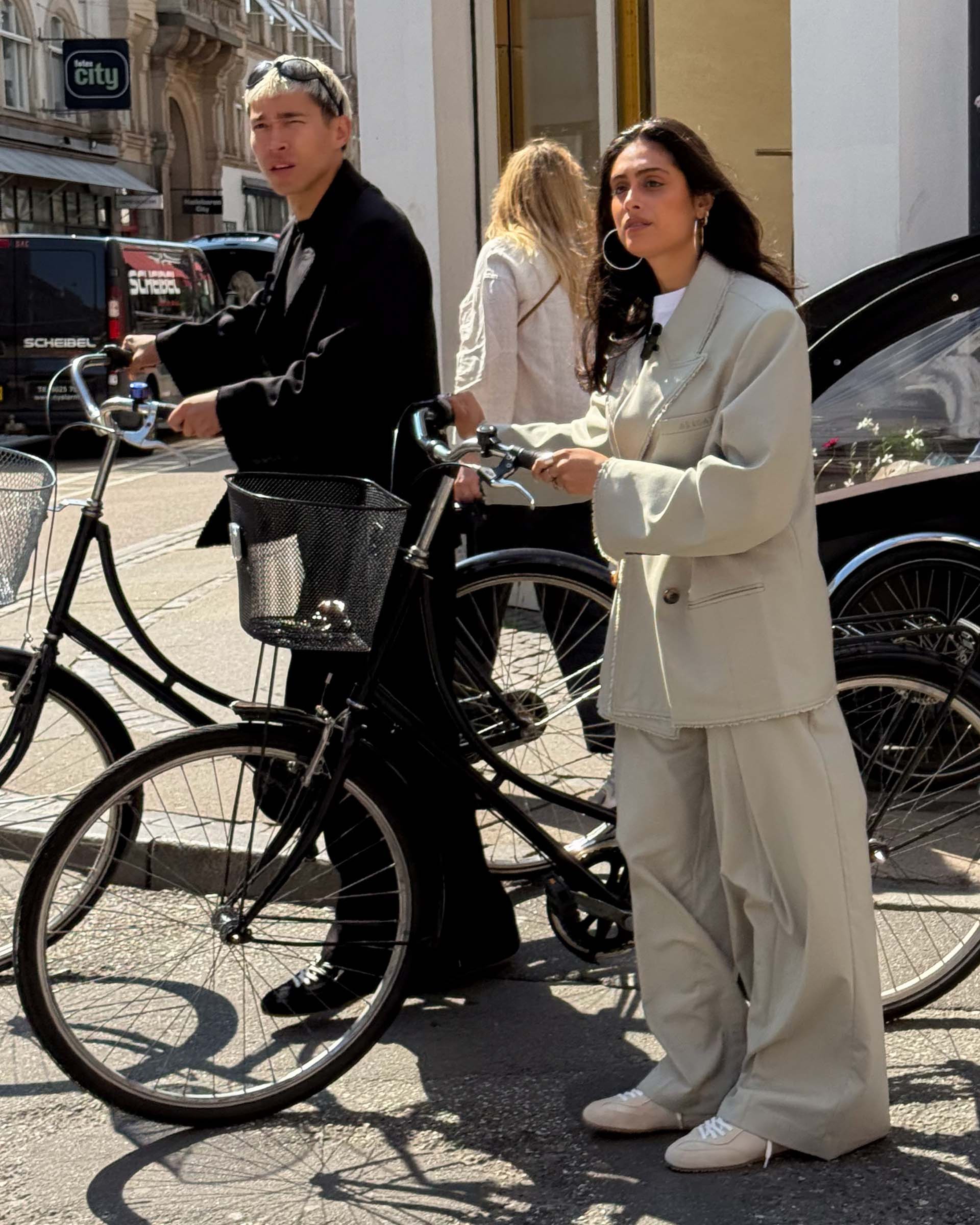 Two people in stylish outfits stand with bicycles on a city street. The woman wears a light-colored suit, and the man is in dark clothing.