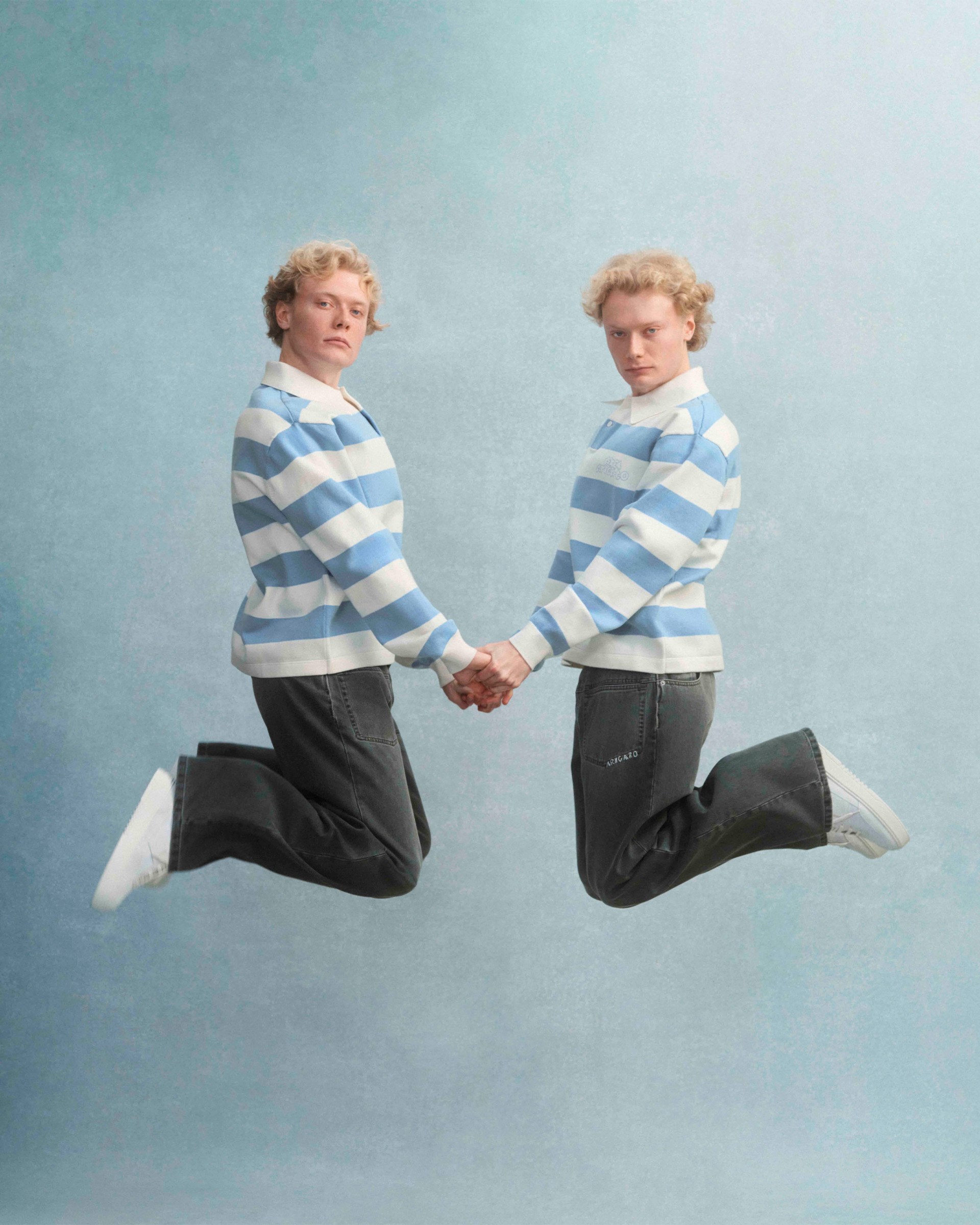 Two people in matching striped sweaters and jeans jump while holding hands against a light blue background.