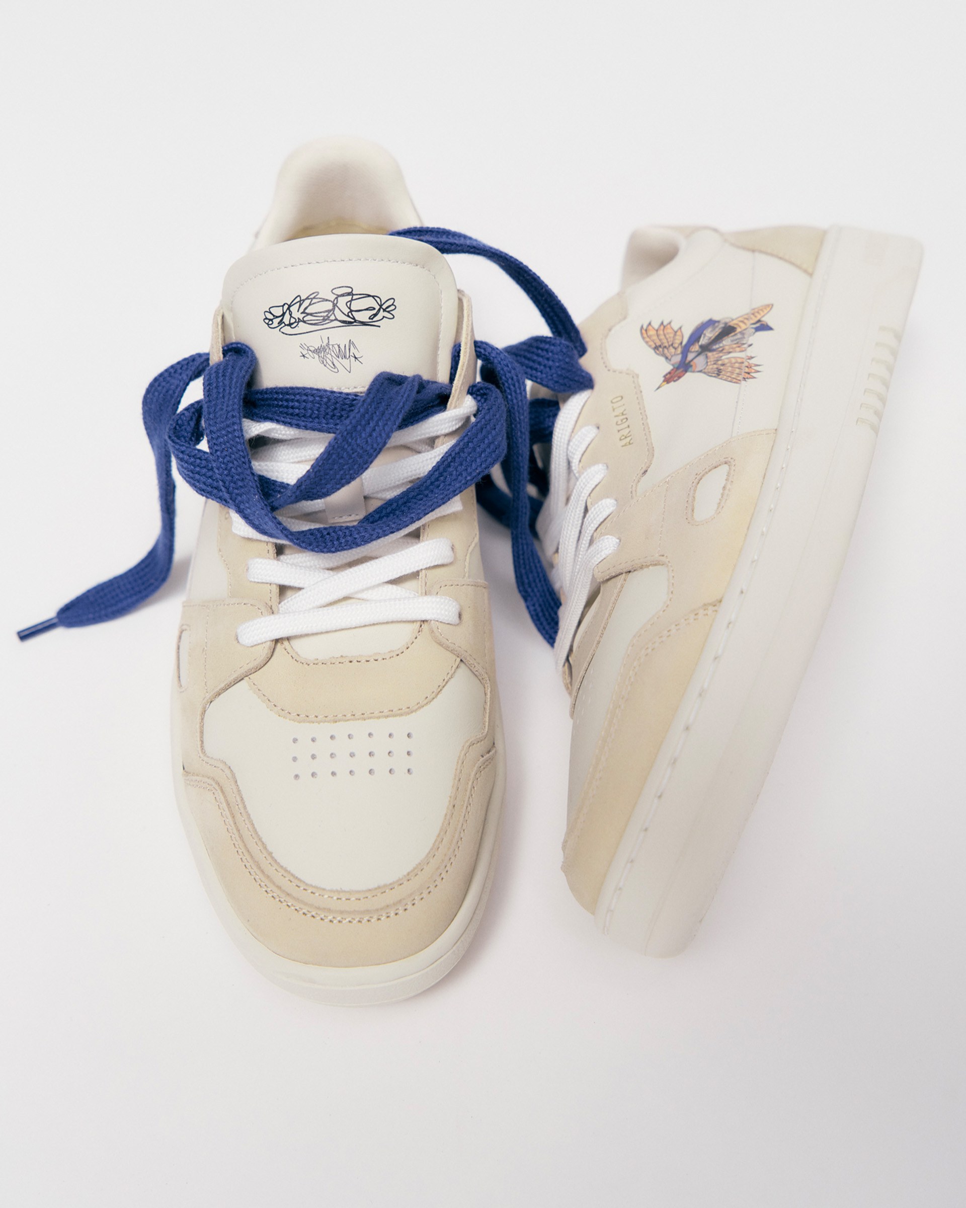 Beige and white sneakers with blue laces; one shoe features a bird design near the heel.