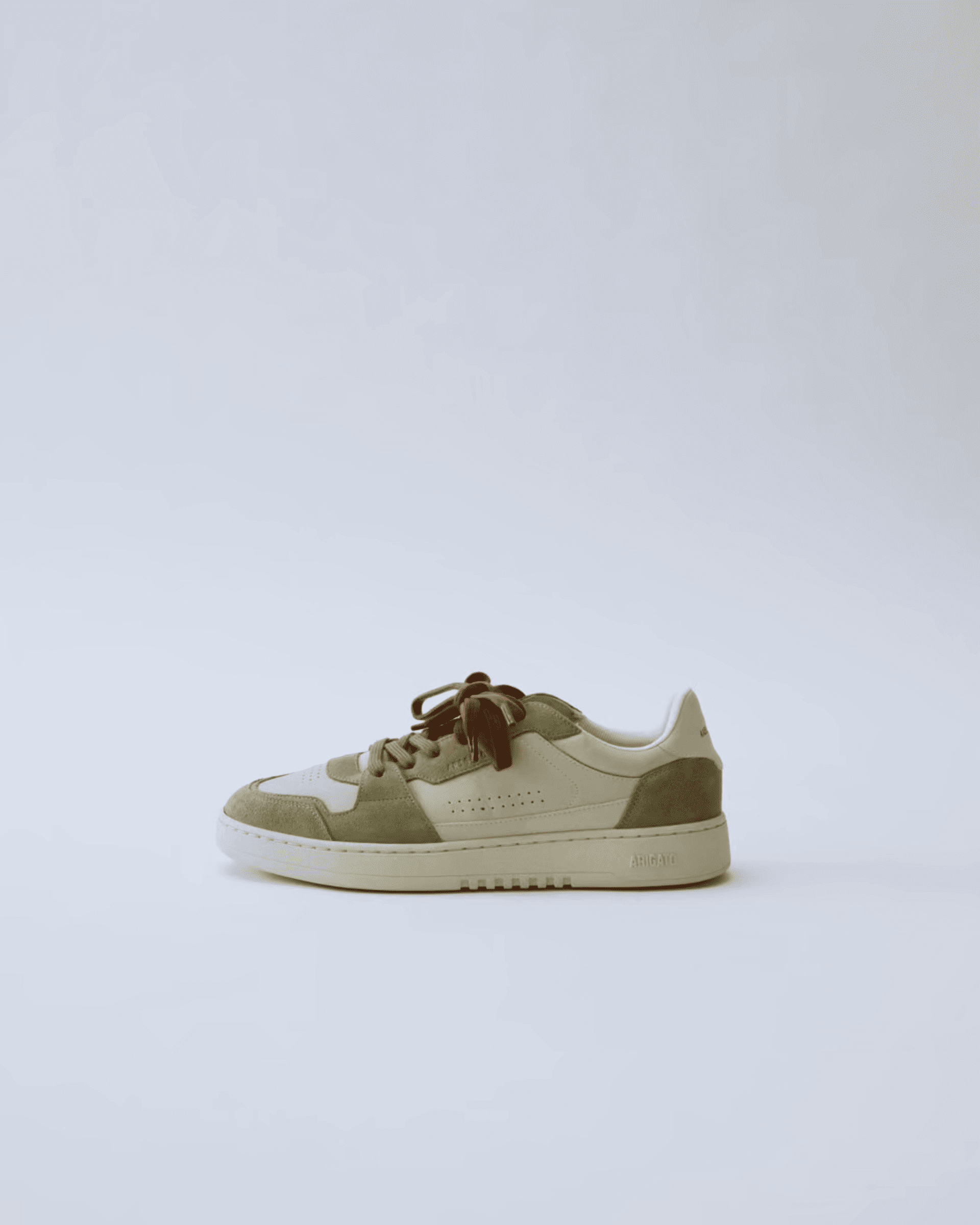Beige and olive green sneaker with brown laces on a plain white background, featuring a perforated design and textured suede elements.