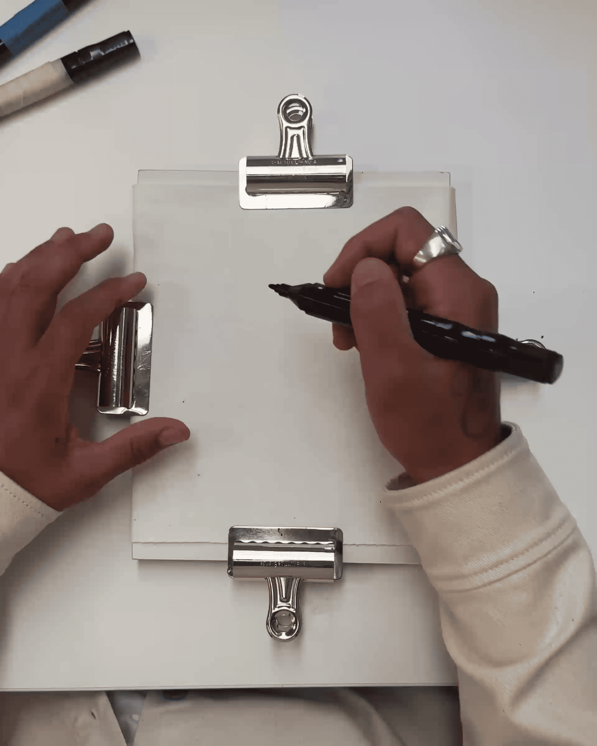 Person writing with a black marker on a clipboard with white paper, wearing a white sleeve and a silver ring.