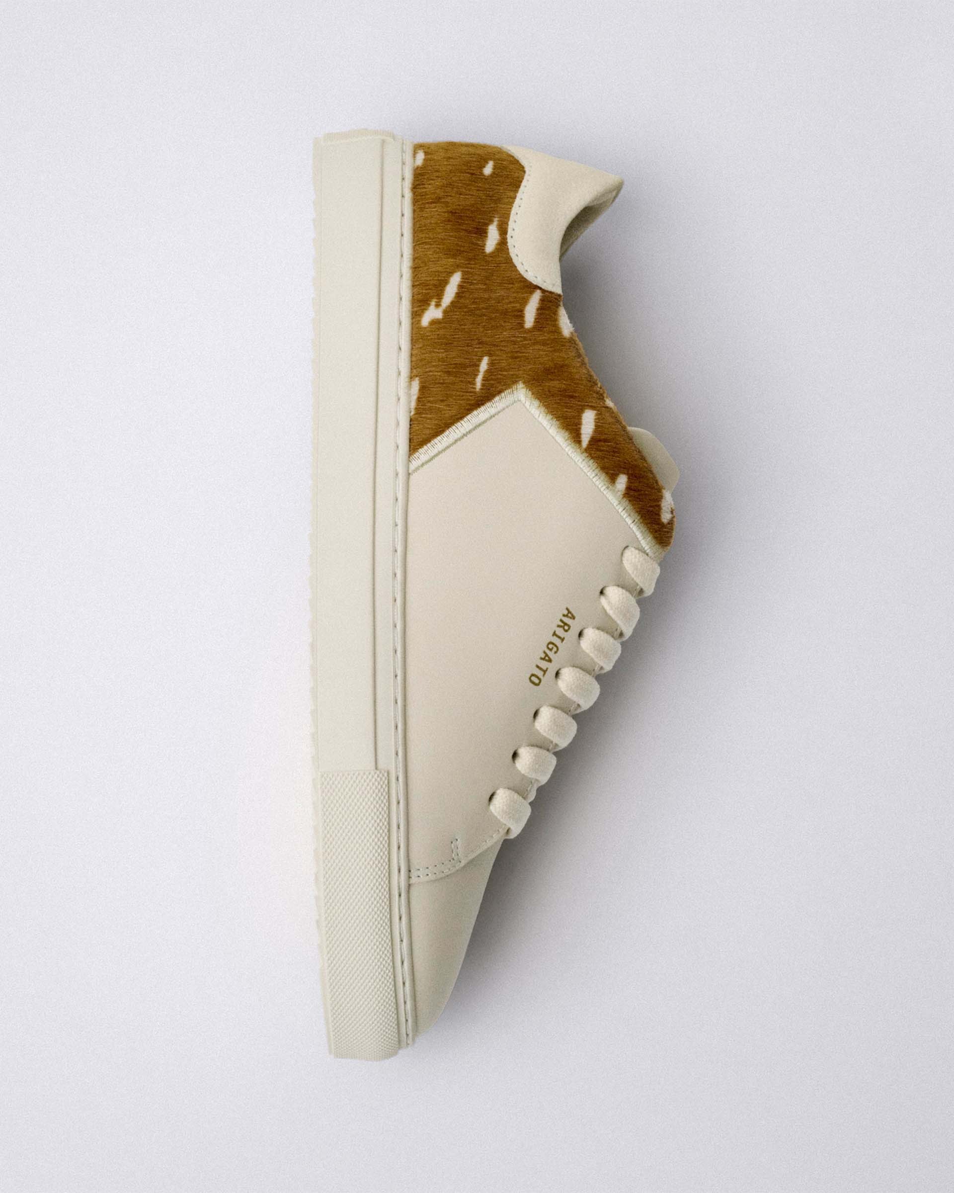 Side view of a stylish sneaker with a white sole and cream leather, featuring a brown and white hair-on panel and embossed branding.