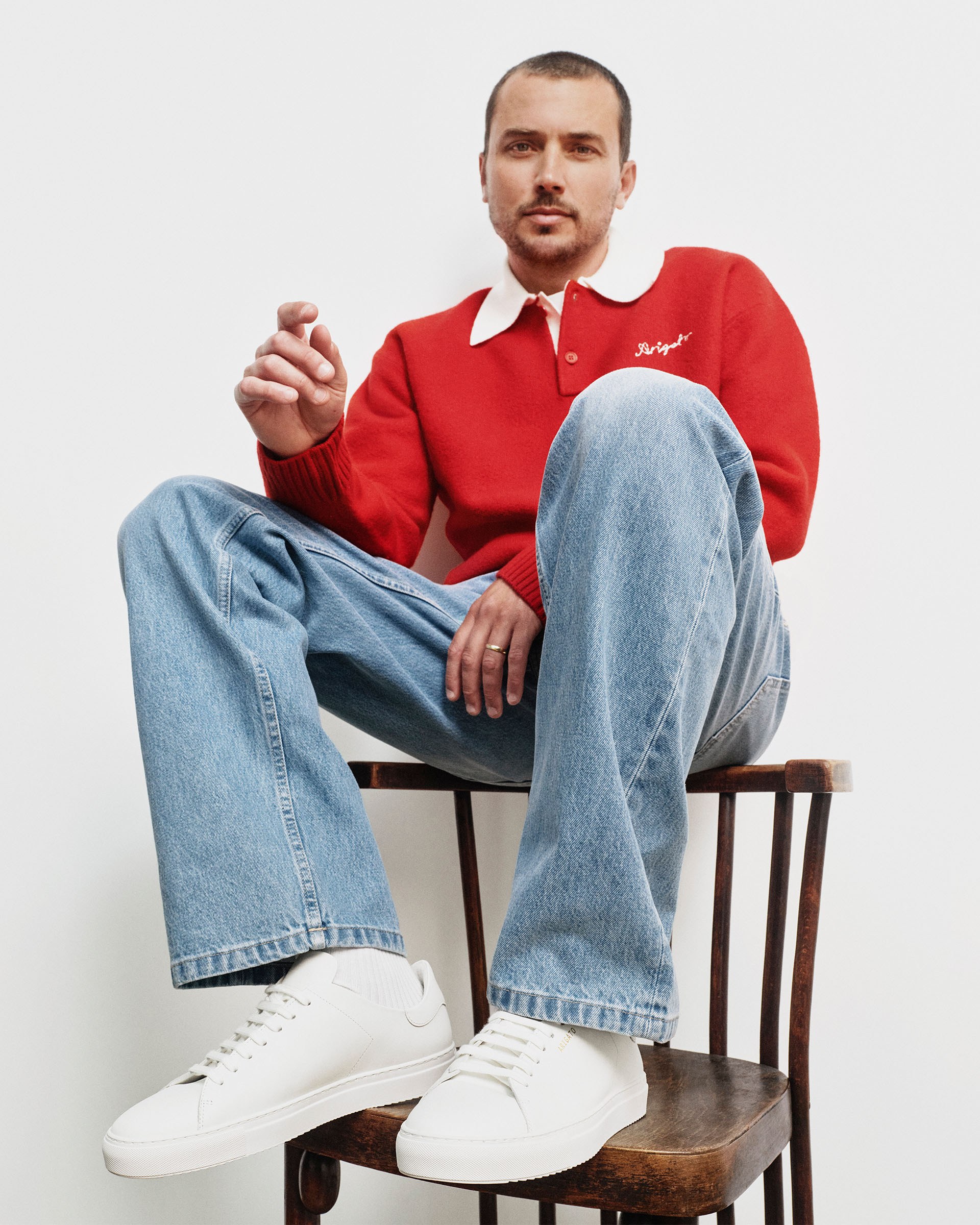 Man seated on a wooden chair, wearing a red sweater, blue jeans, and white sneakers, gazing slightly upward with a relaxed posture.