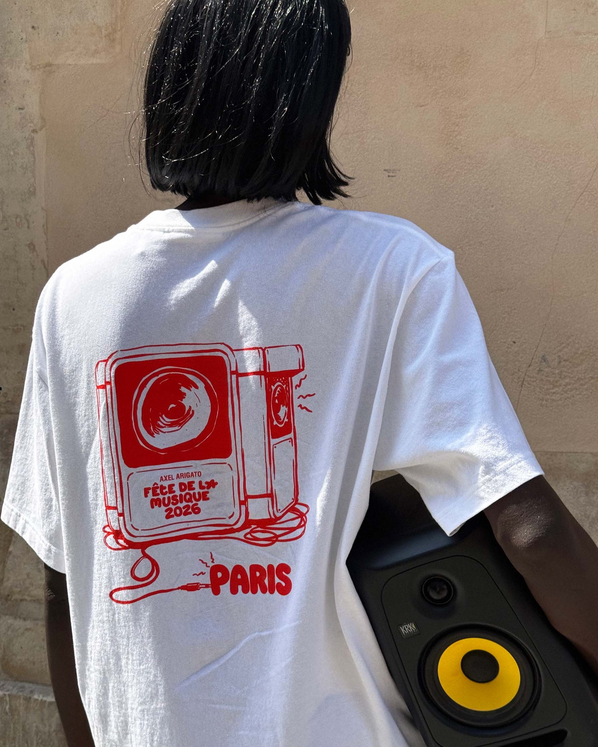 Person in a white T-shirt with a red speaker design and "Fête de la Musique 2026, Paris," carrying a black and yellow speaker.