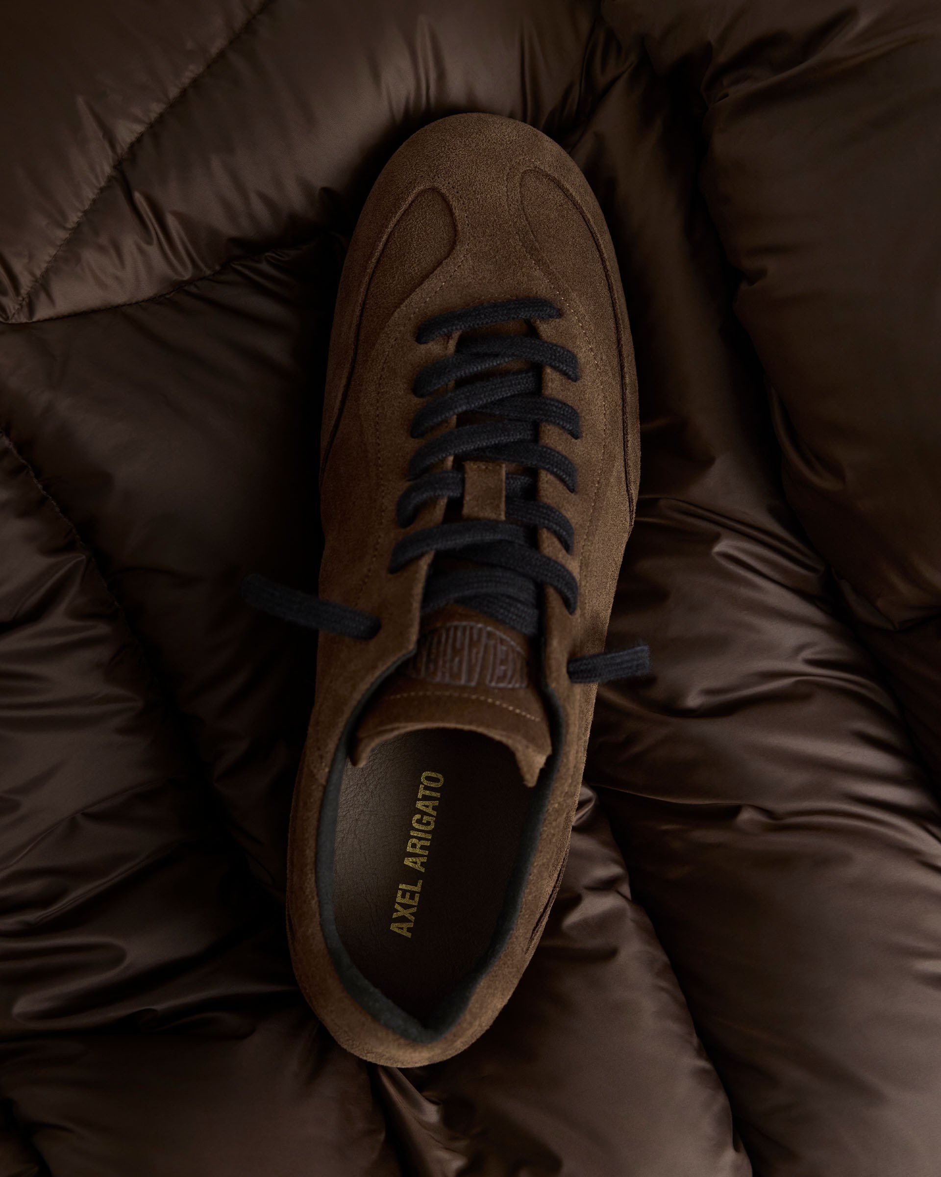 Brown suede sneaker with black laces, labeled "Axel Arigato," placed on a dark, quilted fabric background.