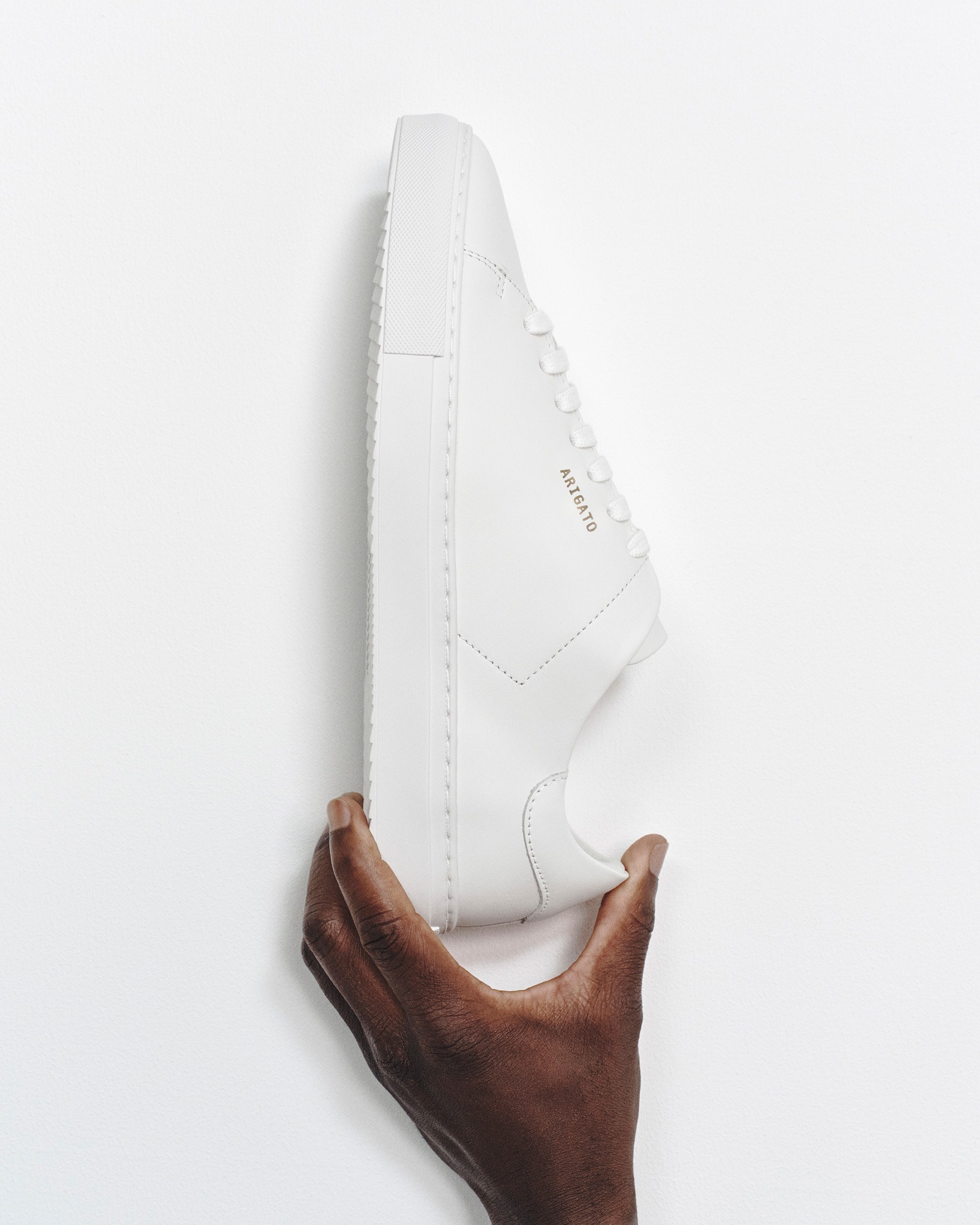 A hand holding a sleek white sneaker with minimal design and gold embossing on the side against a white background.
