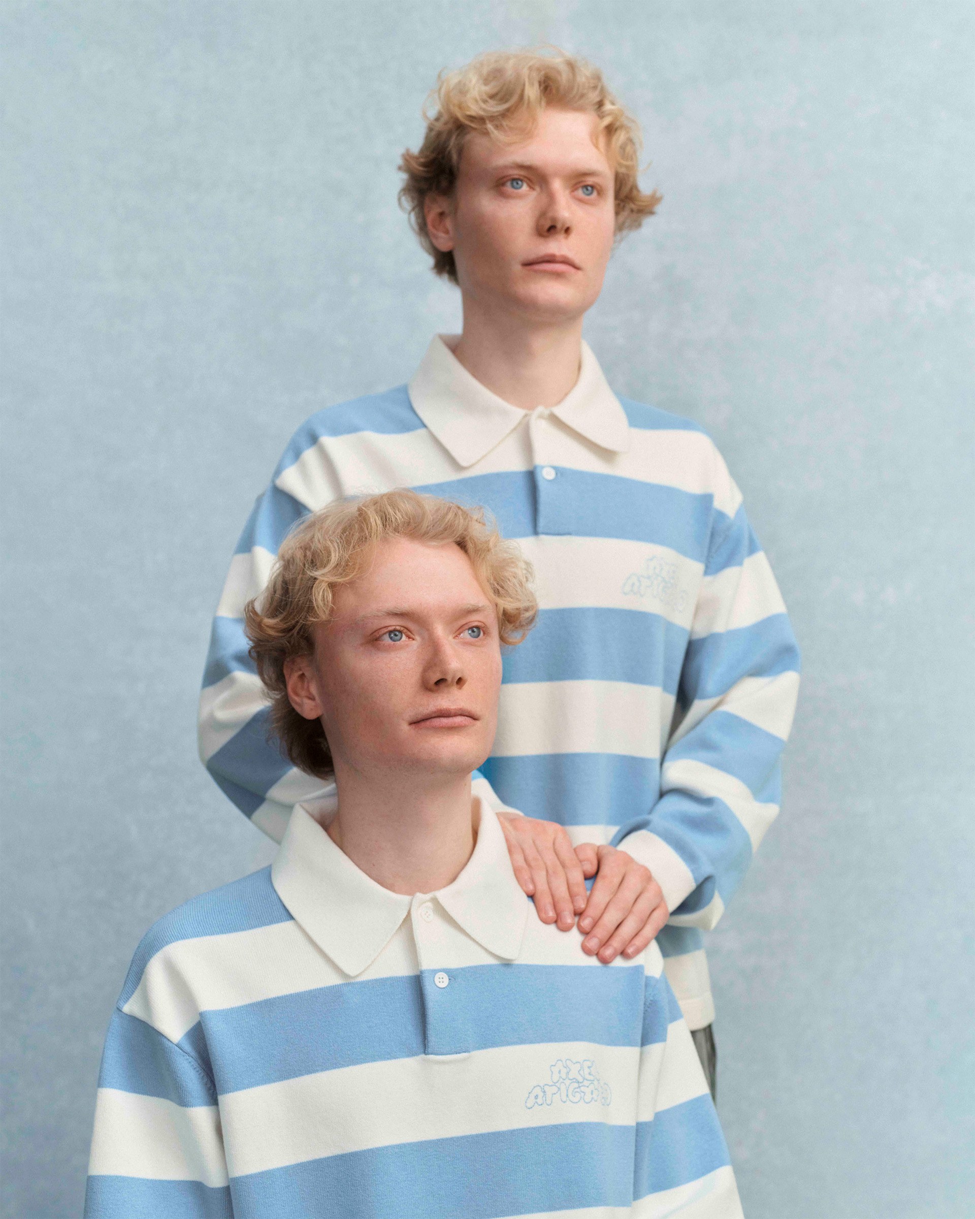 Two individuals with curly blond hair wearing blue and white striped shirts stand against a soft blue background. One rests a hand on the other's shoulder.
